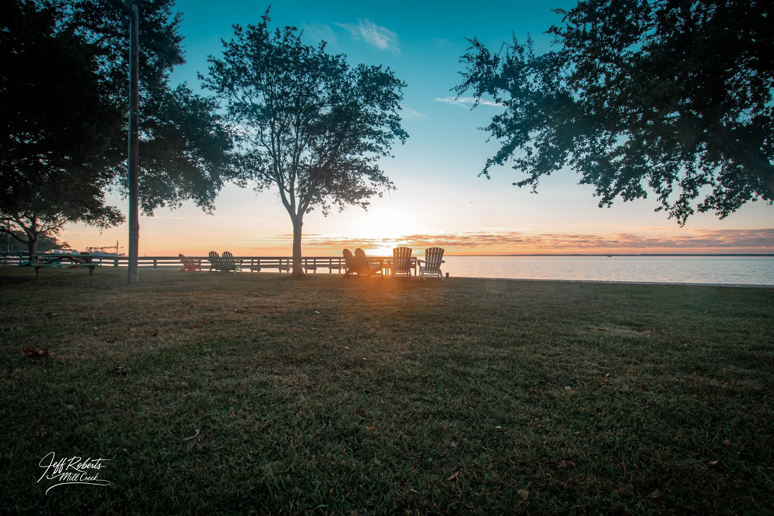 Sunset over a lake viewed from a grassy park with several trees and four Adirondack chairs, two of which are occupied by people, and a wooden fence in the background.