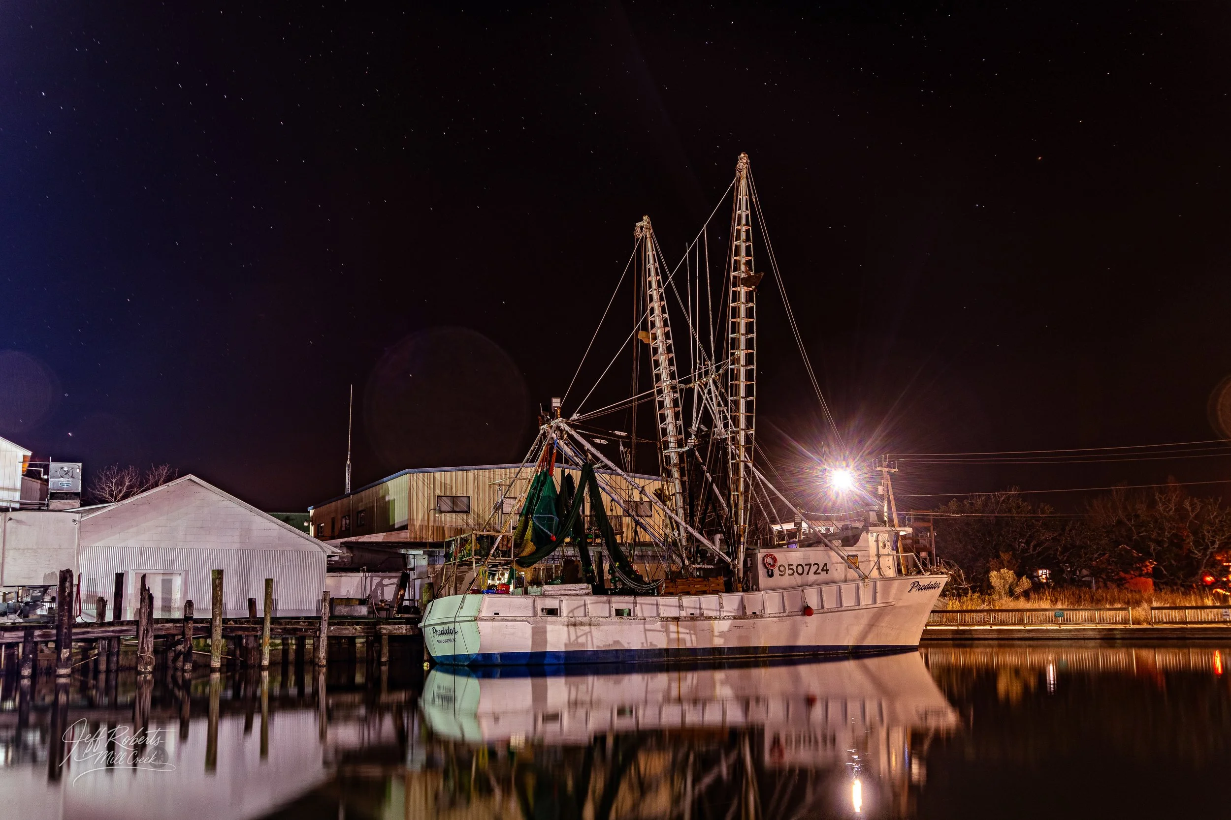 Nighttime scene of a fishing boat named 'Predator' docked at a pier, with starry sky overhead and calm water reflecting the boat and nearby buildings.