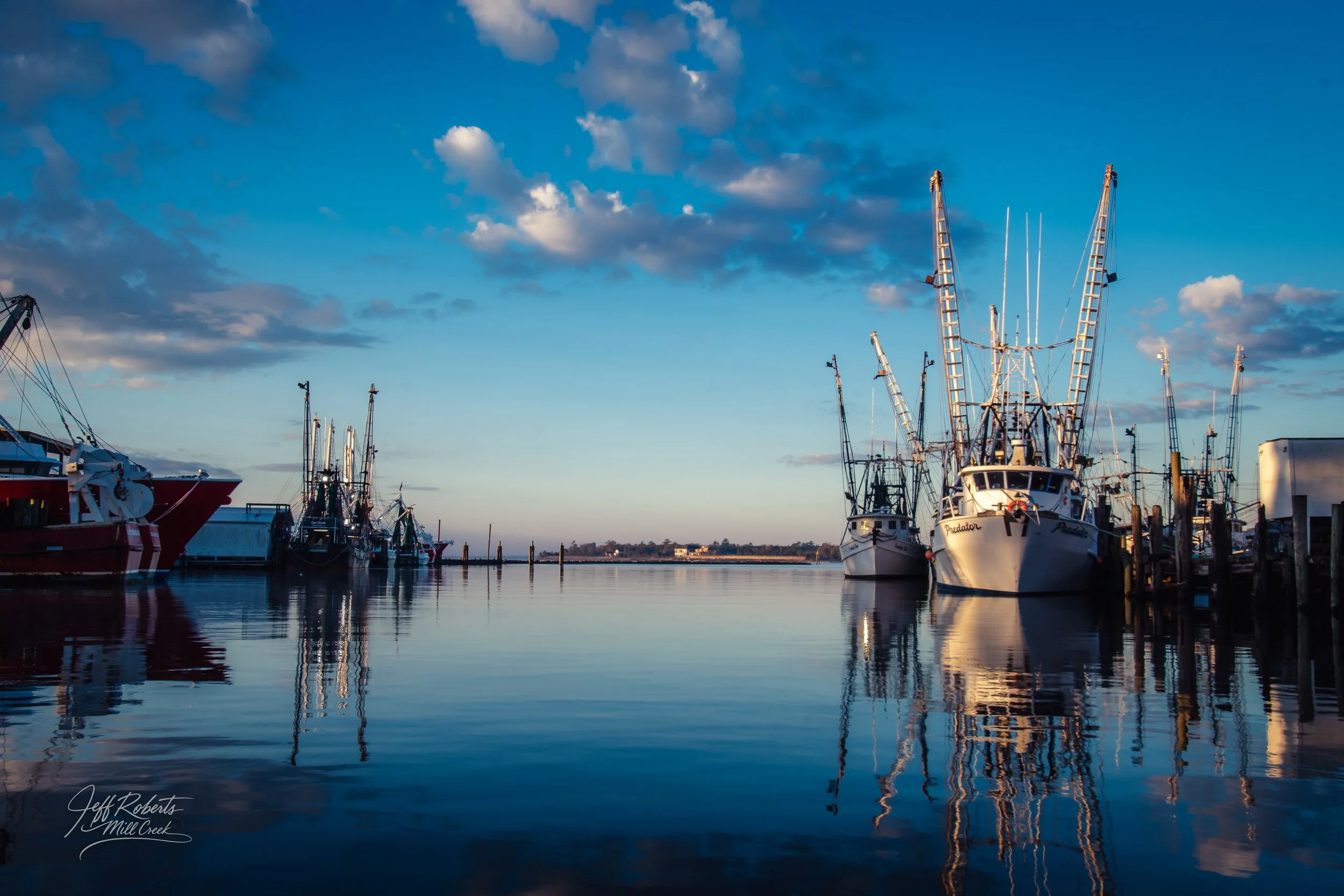 Several fishing boats docked at a marina on a calm body of water with a blue sky and some clouds, taken during sunset or sunrise.