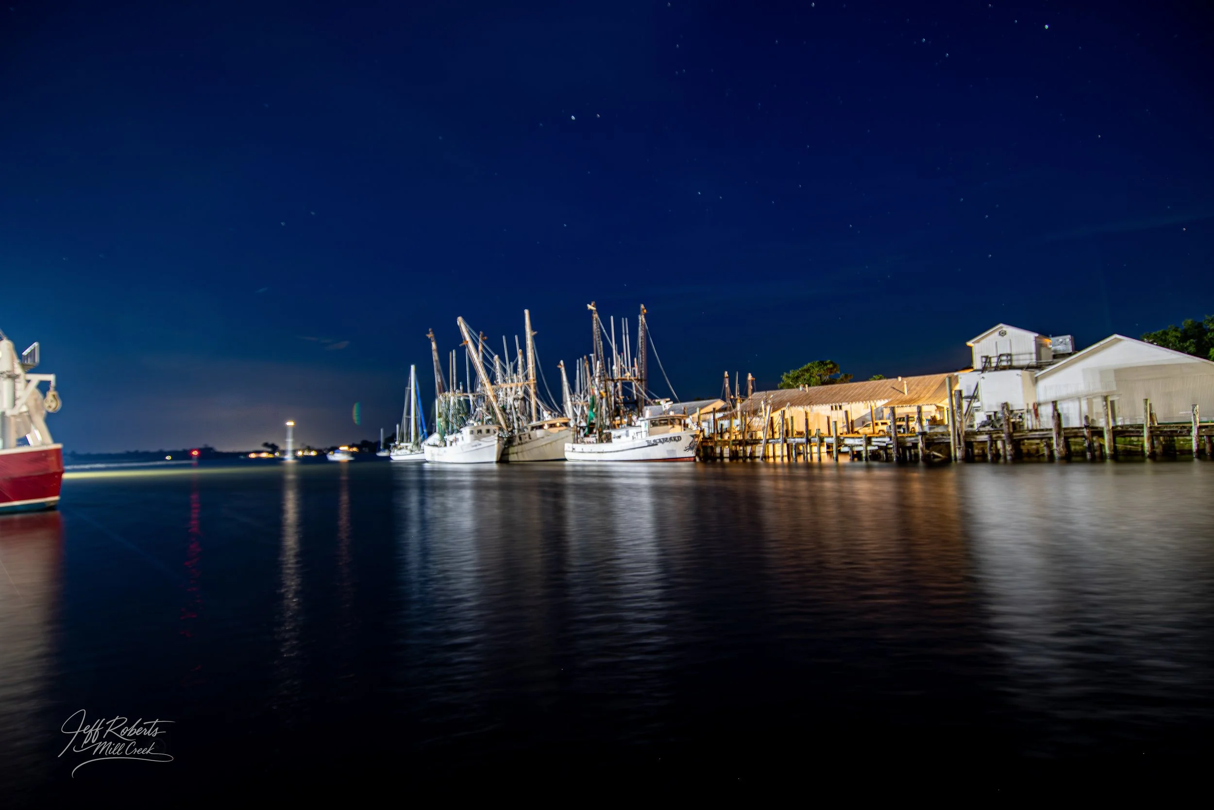 Nighttime view of boats docked at a marina, with buildings and a starry sky in the background.