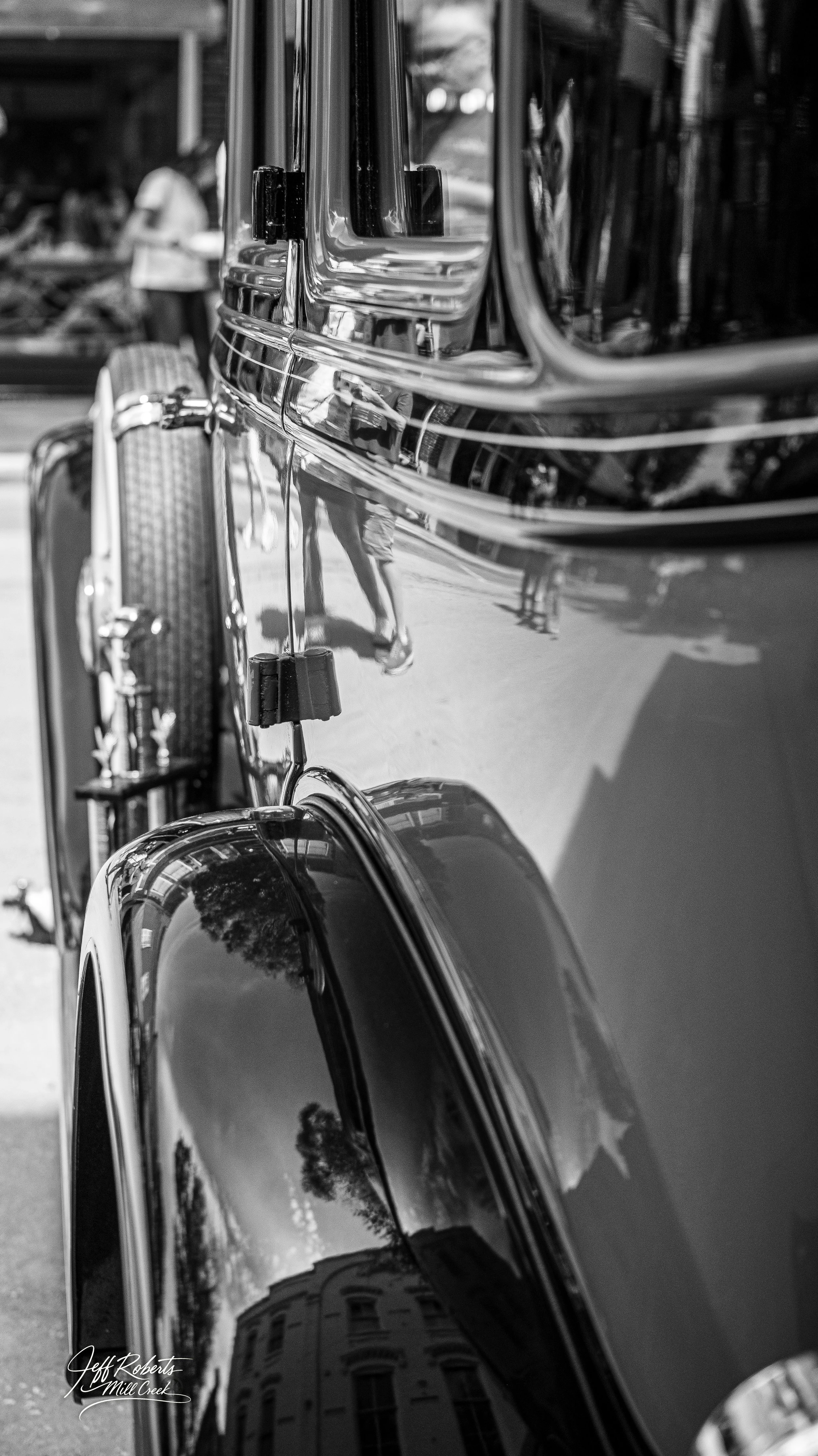 Close-up of a vintage car's chrome and black paint with reflections of trees and buildings.