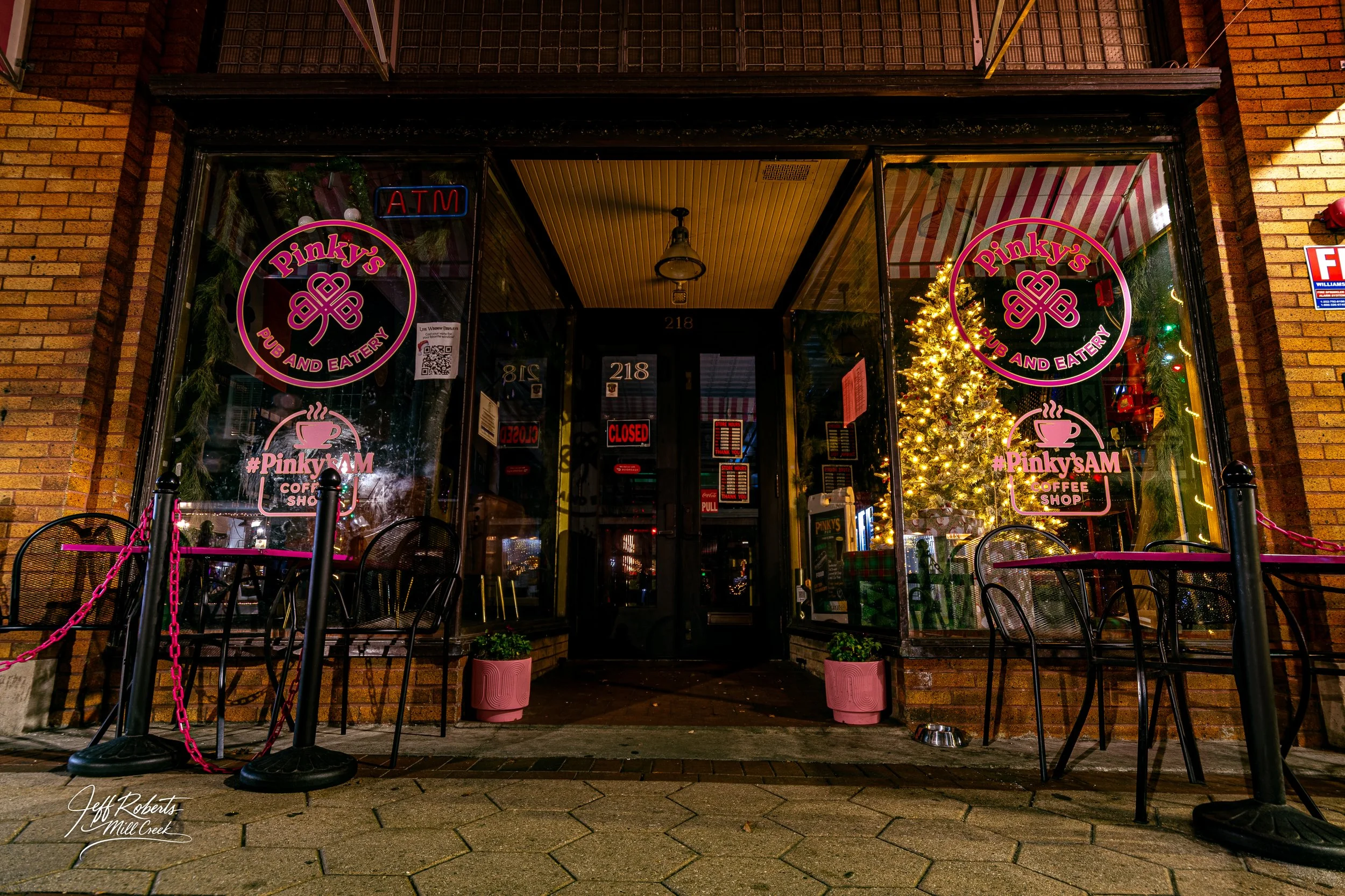 Nighttime exterior of Pinky's Pub and Eatery with Christmas tree inside, illuminated through large front windows, and outdoor seating area with black chairs and pink chains.