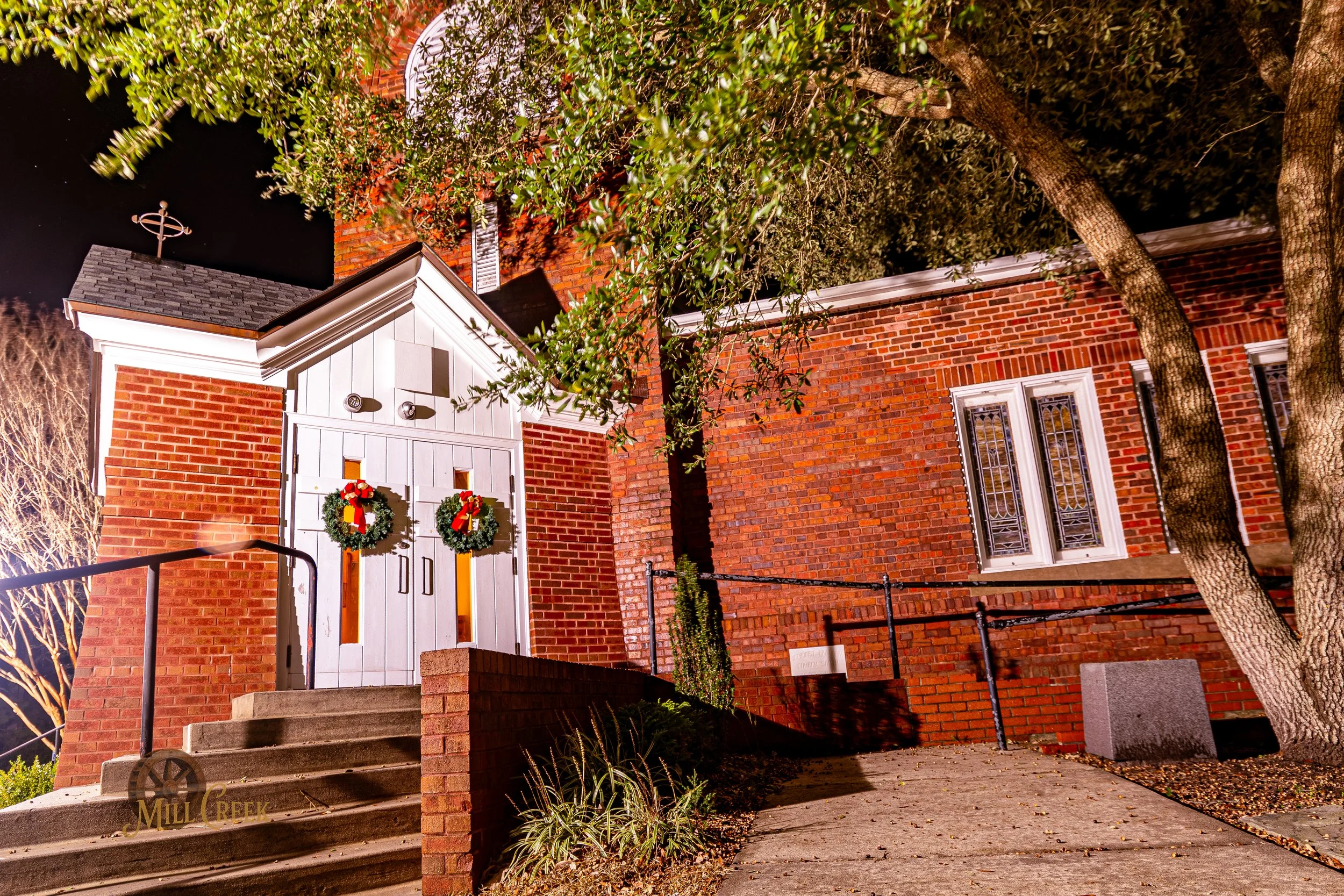 Red brick building decorated with two Christmas wreaths on the white door, outdoor stairs, and trees at night.