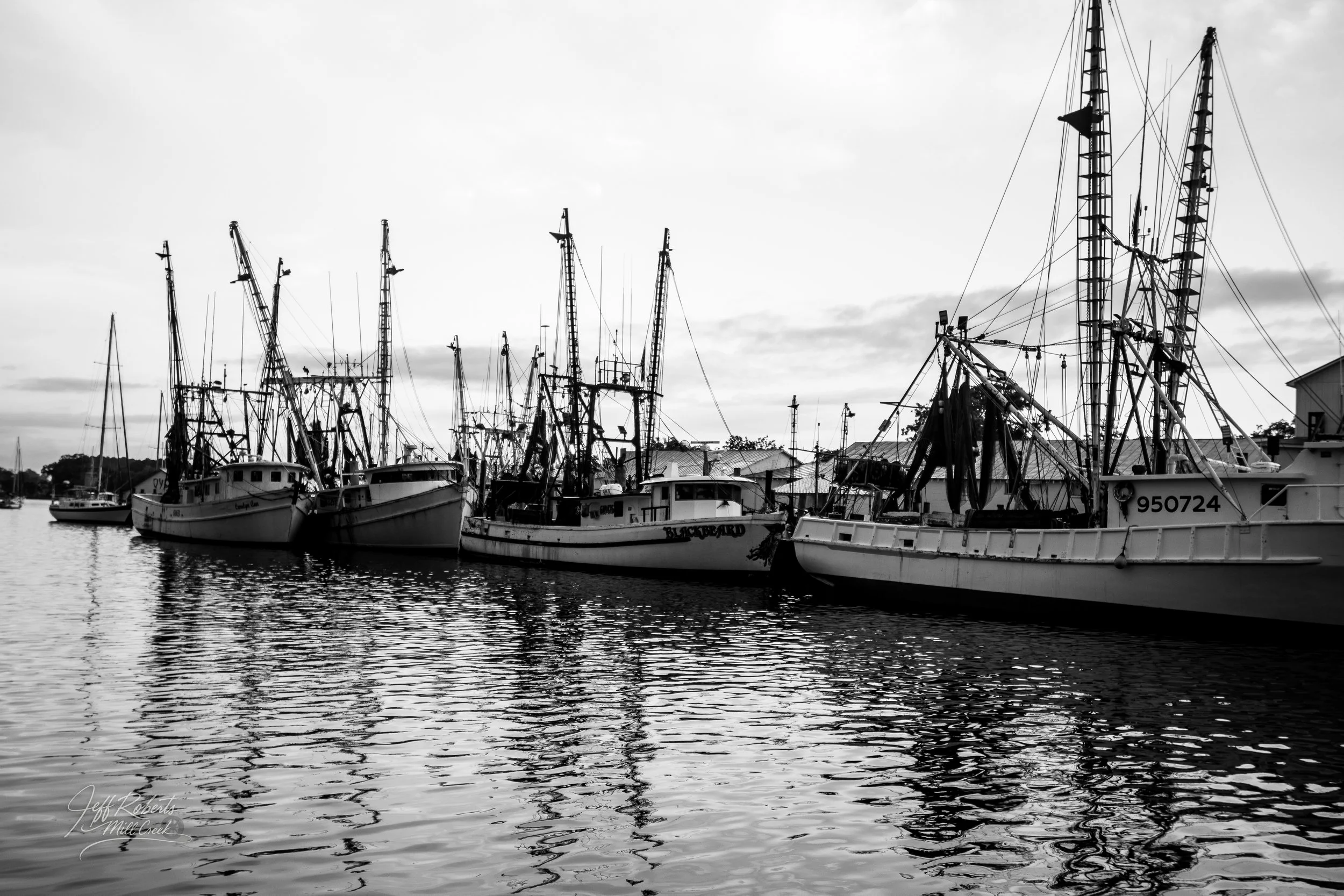 Black and white photograph of fishing boats docked at a marina, with reflections on the water.