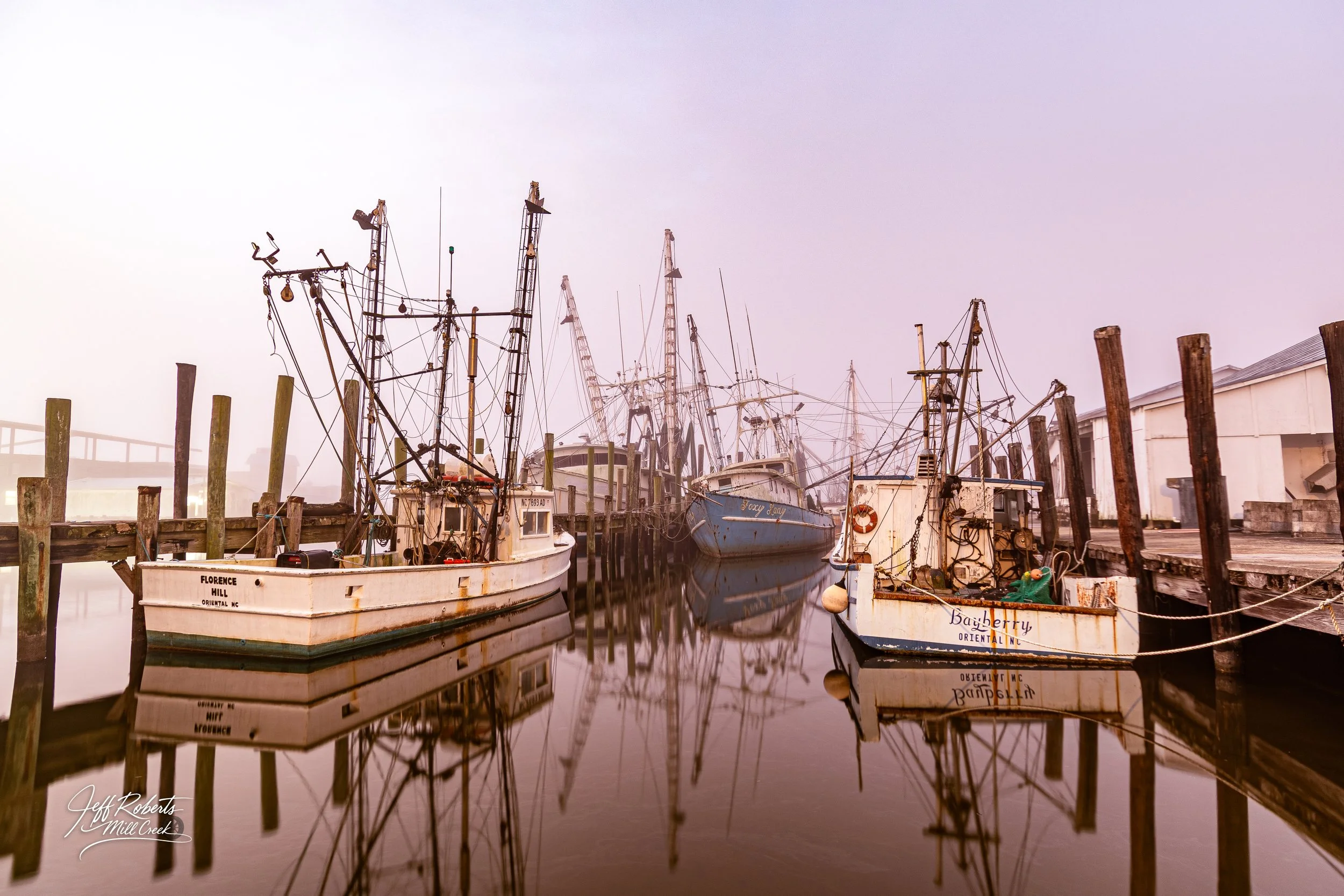 Foggy morning at a dock with several fishing boats moored, their reflections visible in the calm water, with weathered wooden posts and a white building in the background.