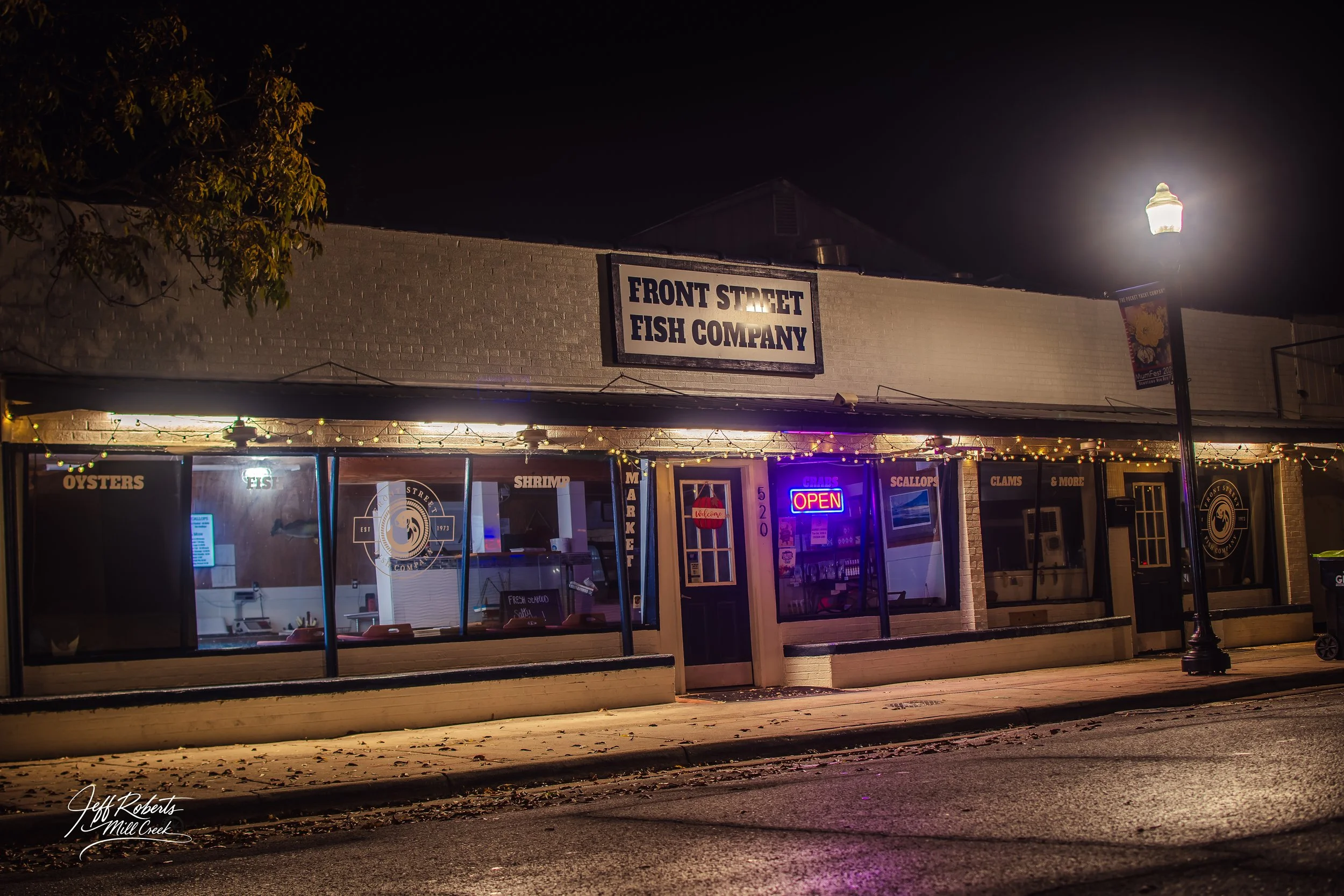 Night view of Front Street Fish Company with a sign, windows displaying signs for oysters, fish, shrimp, scallops, clams, and more, and a neon "Open" sign.