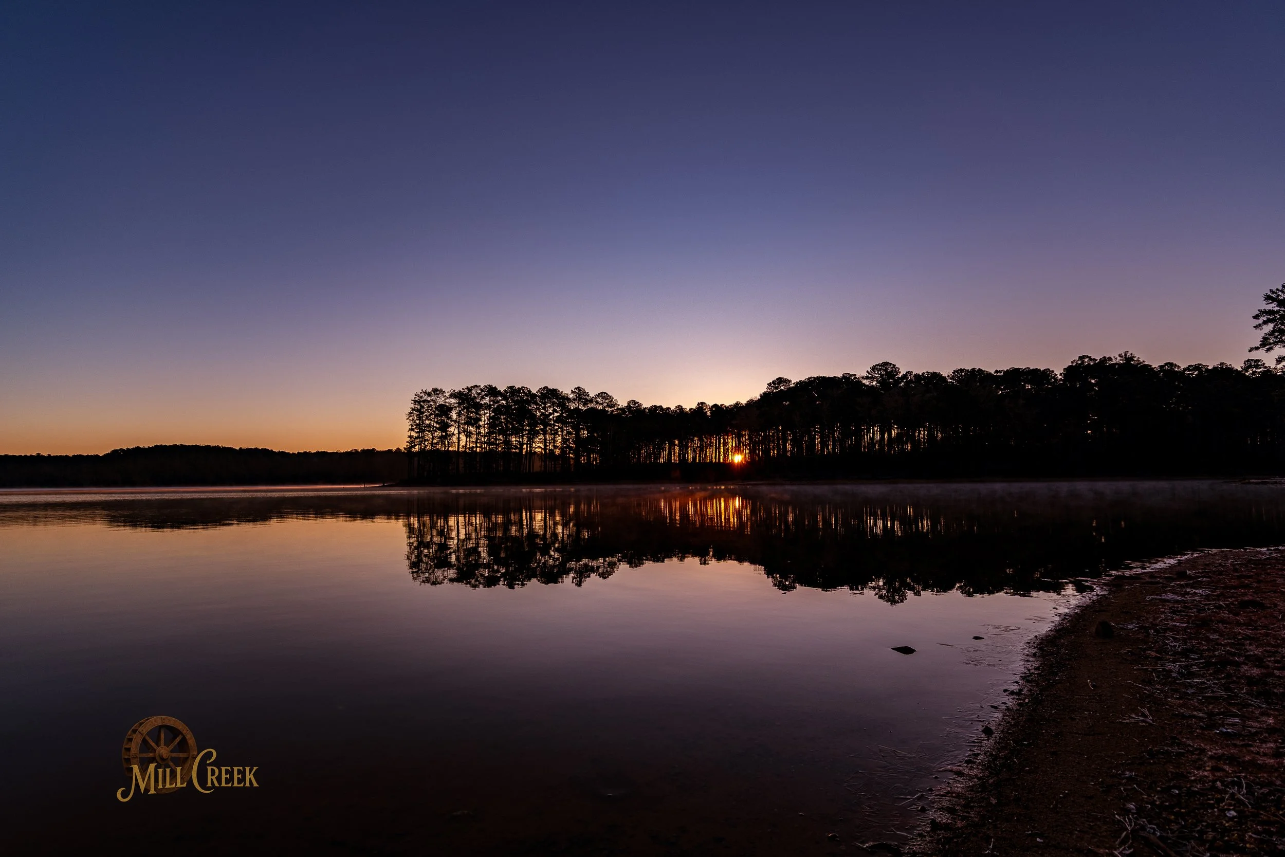 Sunset over a calm lake with trees on the horizon and their reflection in the water, with a sandy shoreline in the foreground. Mill Creek logo in the bottom left corner.