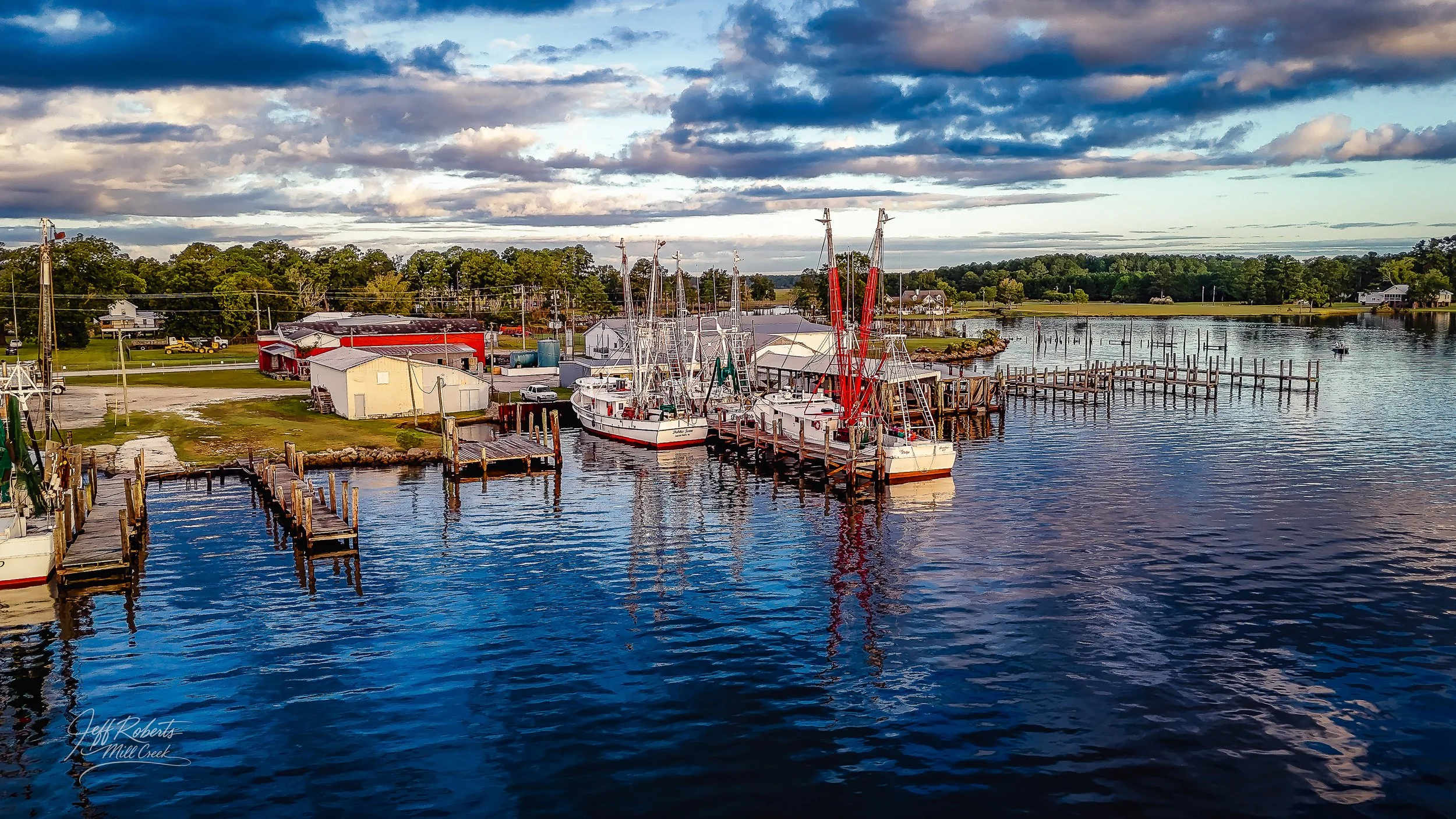 Boats docked at a marina on a calm body of water under a partly cloudy sky, with trees and buildings in the background.
