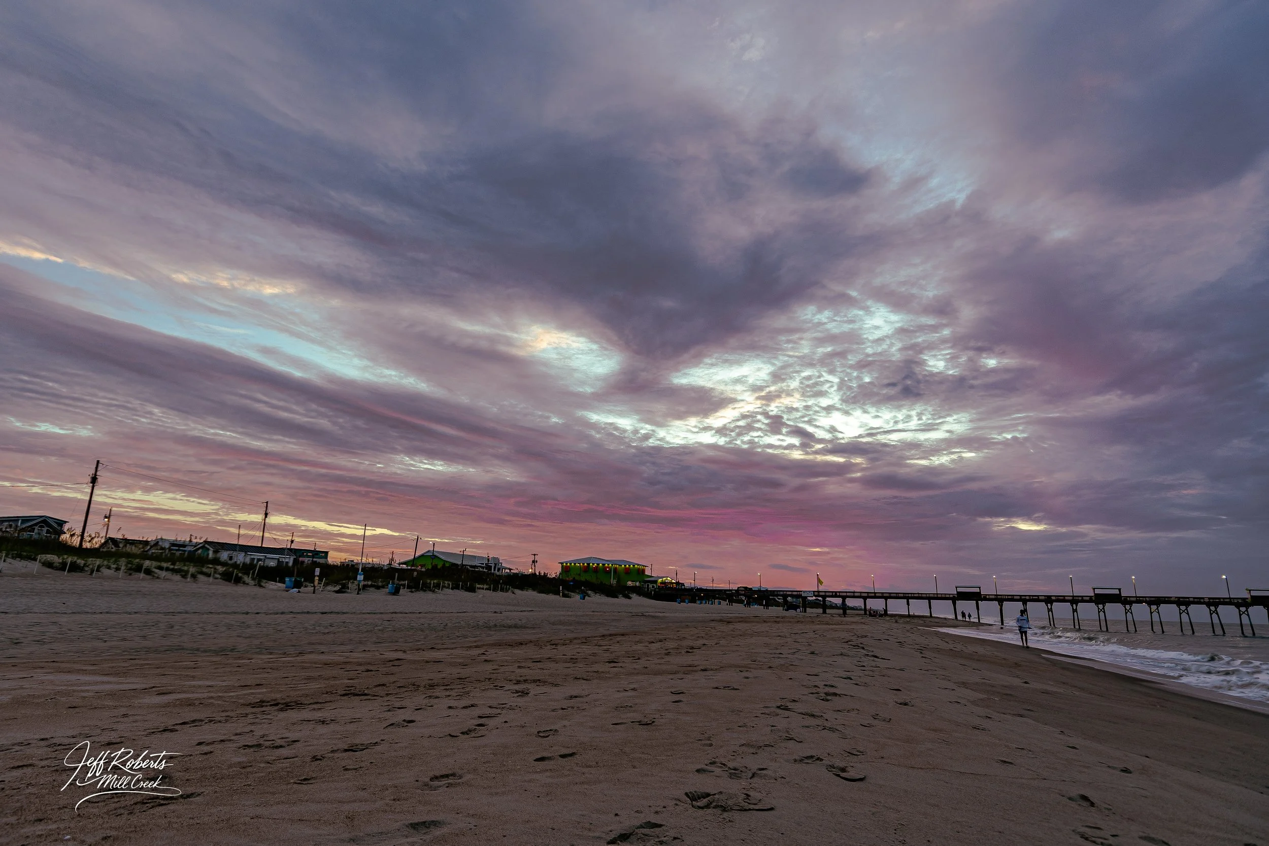 Sunset over a sandy beach with footprints, pier extending into the ocean, and colorful sky with clouds.