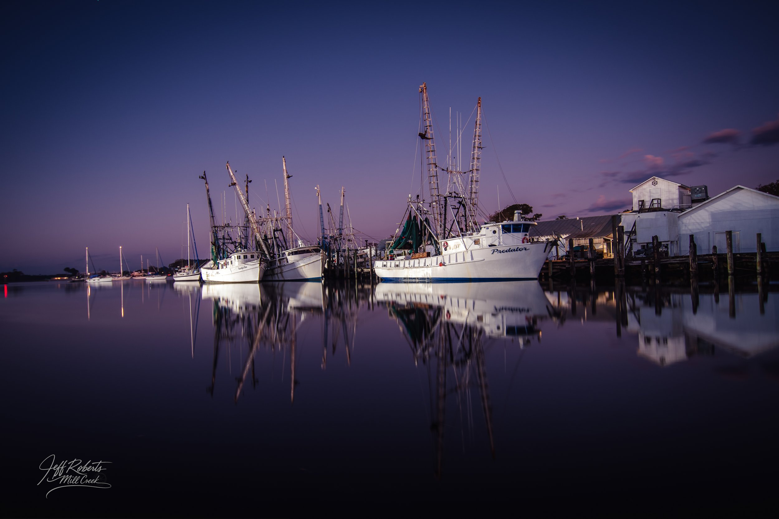 A calm harbor at dusk with several sailboats docked, their masts and hulls reflected in the still water, and a sky transitioning from deep blue to purple.