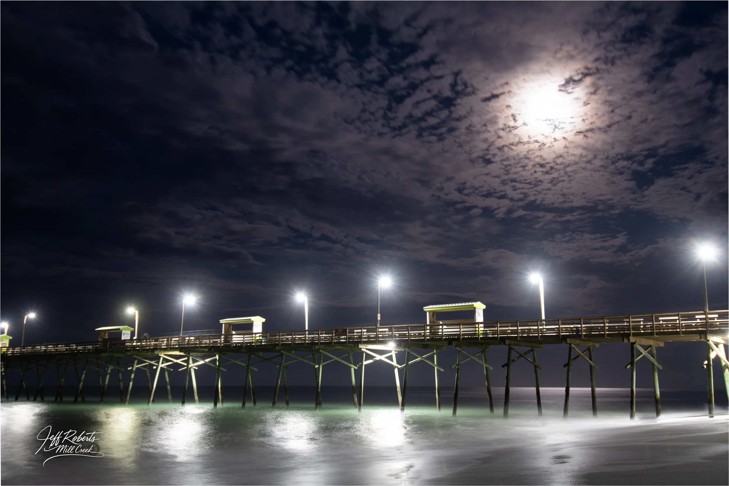 Nighttime view of a wooden pier extending over the ocean, illuminated by bright lights; the moon and partially cloudy sky are visible overhead.