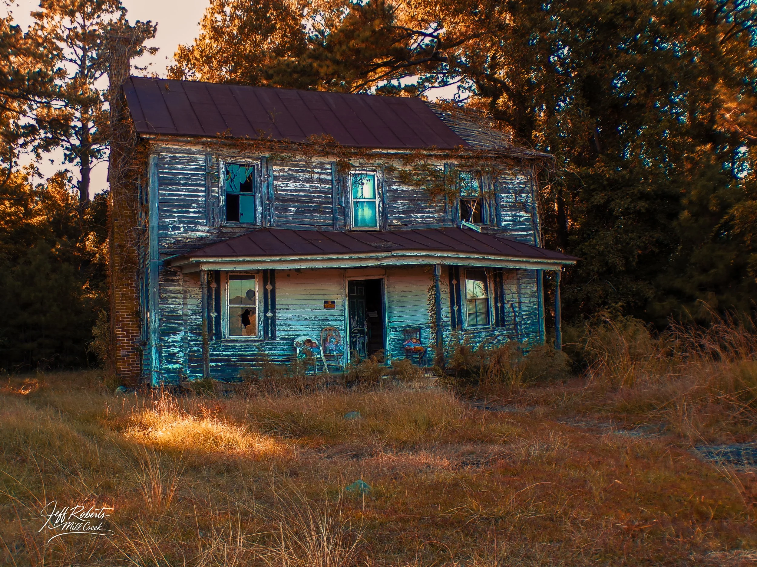 An old, abandoned two-story house with peeling blue paint, broken windows, and a rusted metal roof, surrounded by tall grass and trees during sunset.