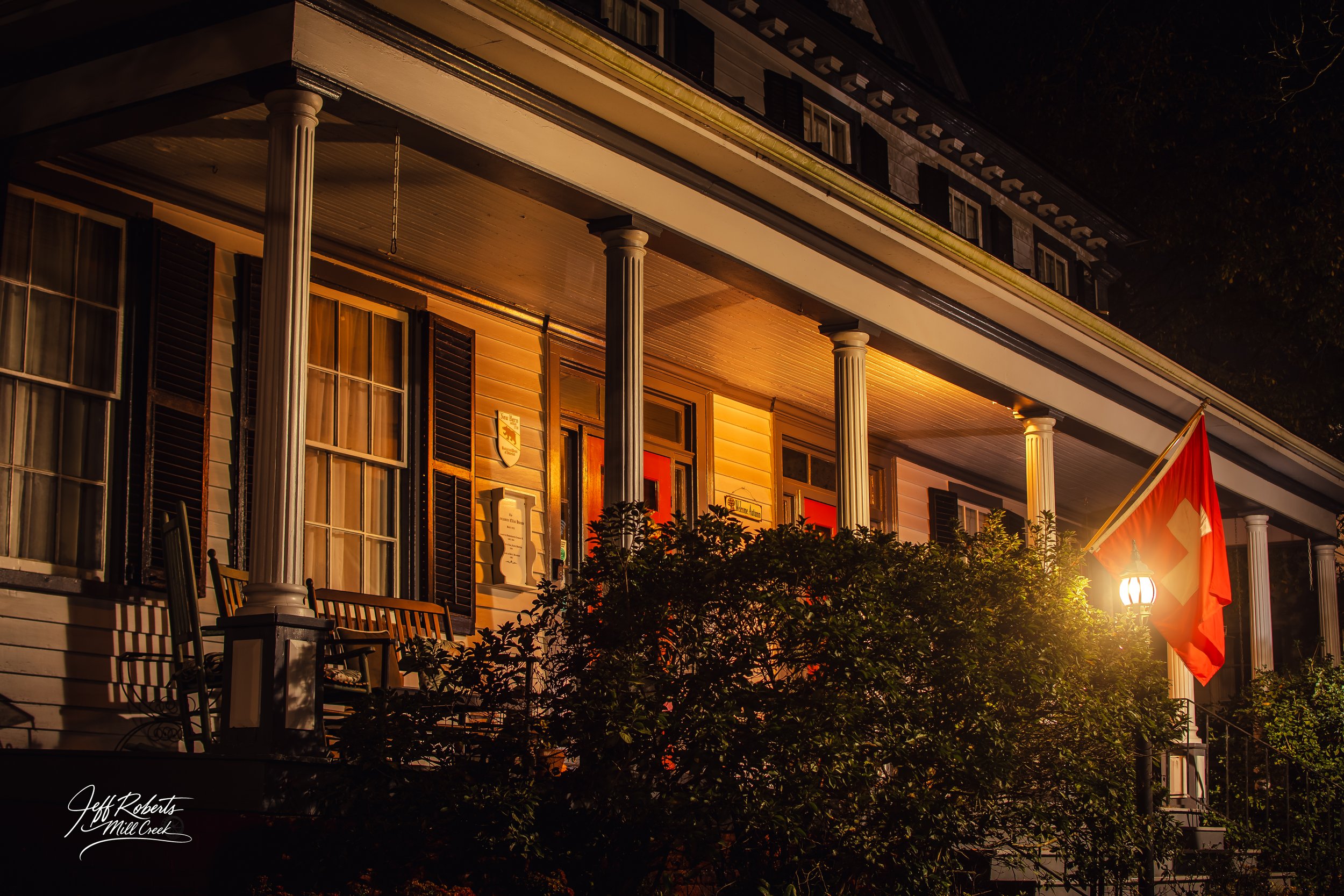 A colonial-style house illuminated at night with a porch supported by white columns, decorated with a Swiss flag, surrounded by bushes, and warm lighting from lanterns.
