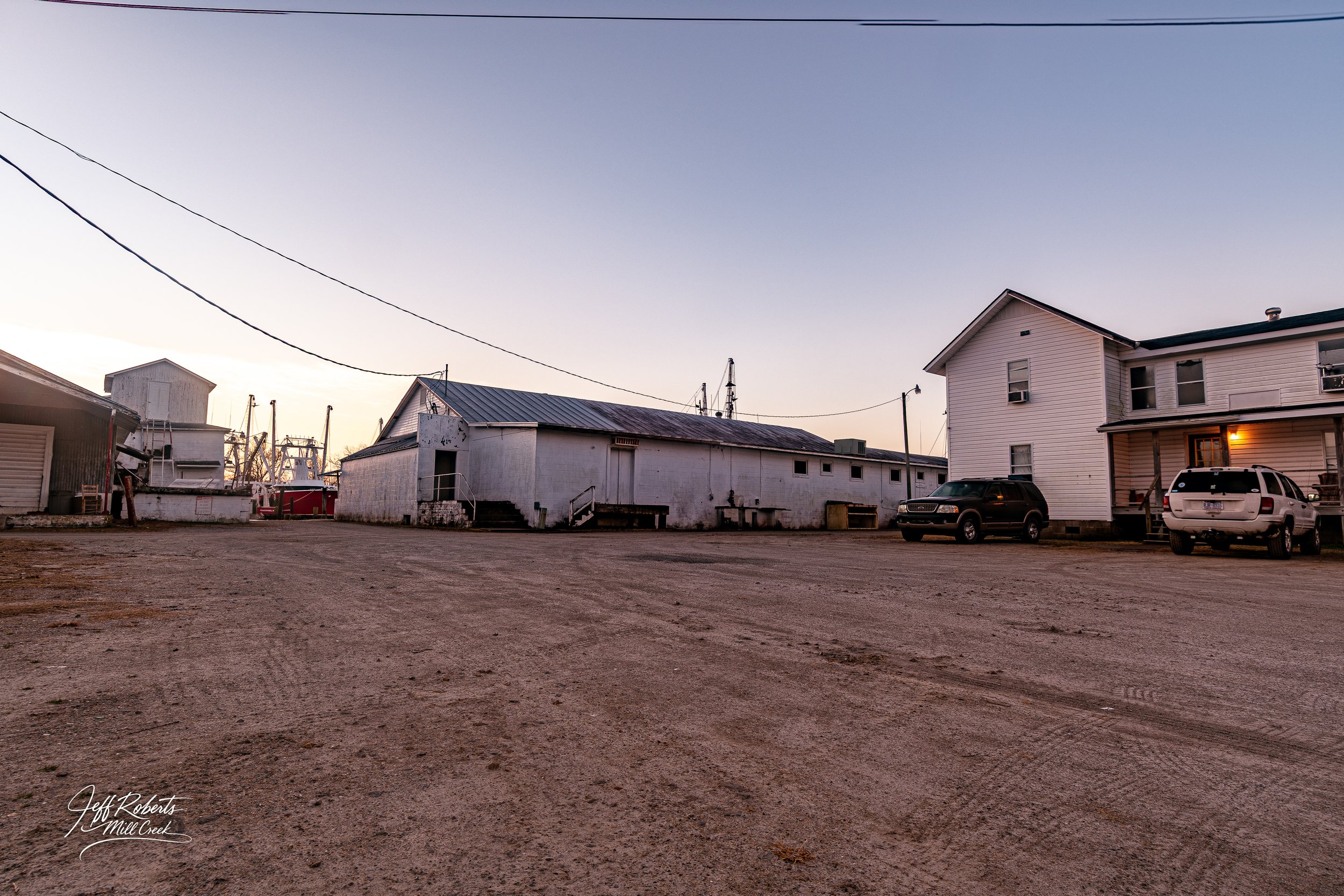 Dirt lot in front of small buildings and houses at sunset, with power lines overhead and parked cars visible on the right.