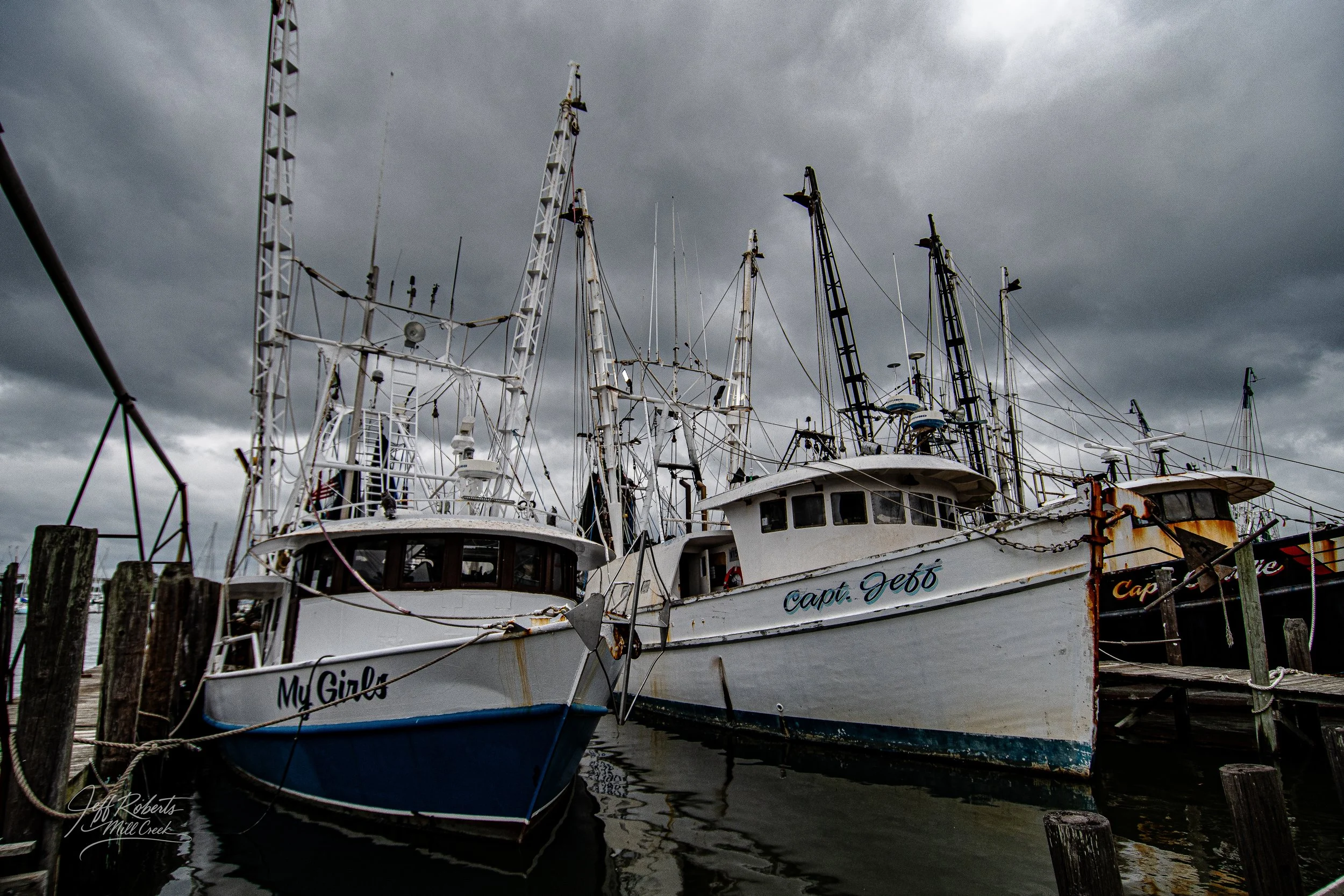 Several fishing boats docked at a marina under dark, cloudy skies. The boats are tied to wooden posts and have nameplates visible, with one reading 'My Girl' and another 'Capt. Jef6.'