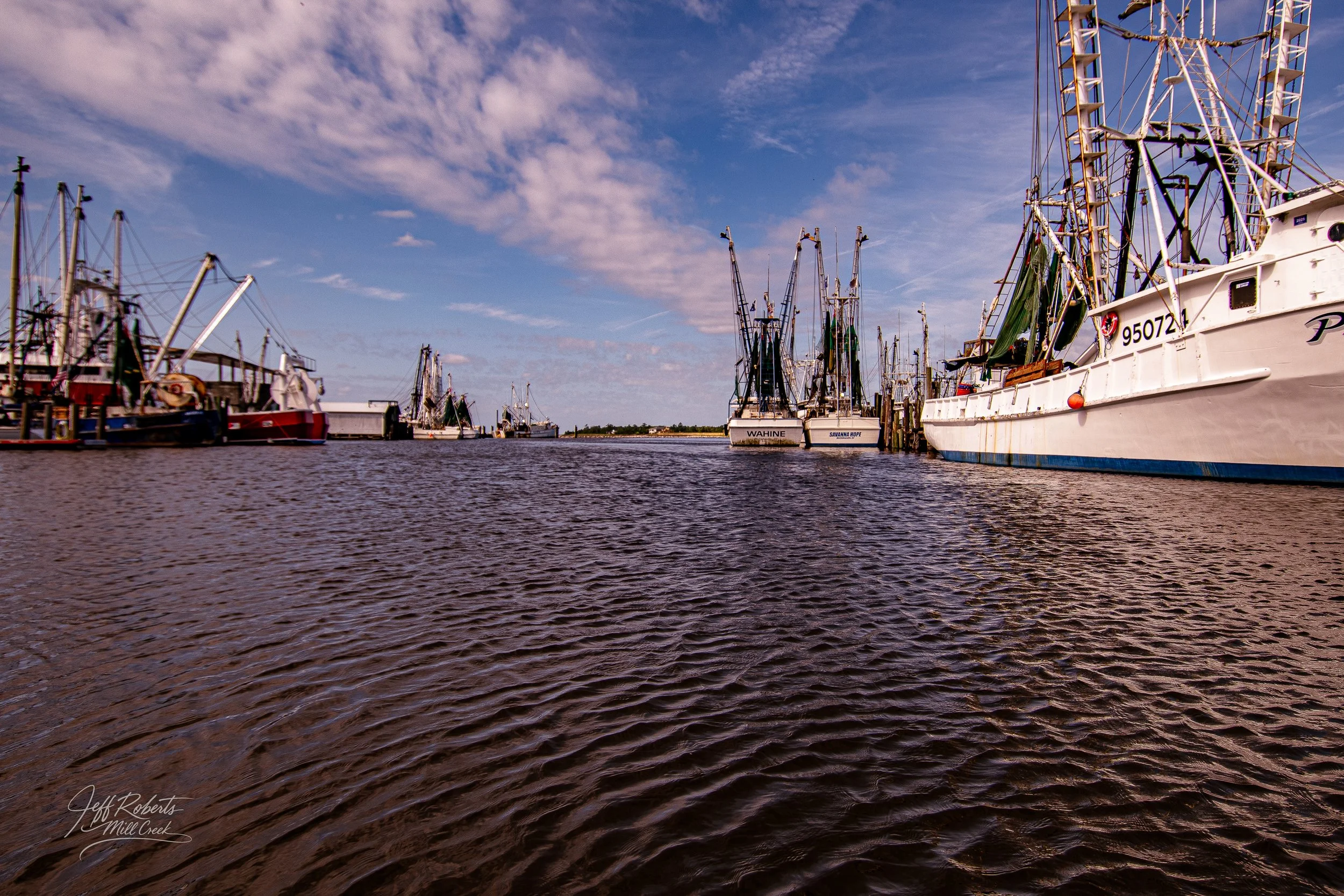 View of a marina with several fishing boats docked on both sides of a calm body of water, under a partly cloudy sky.
