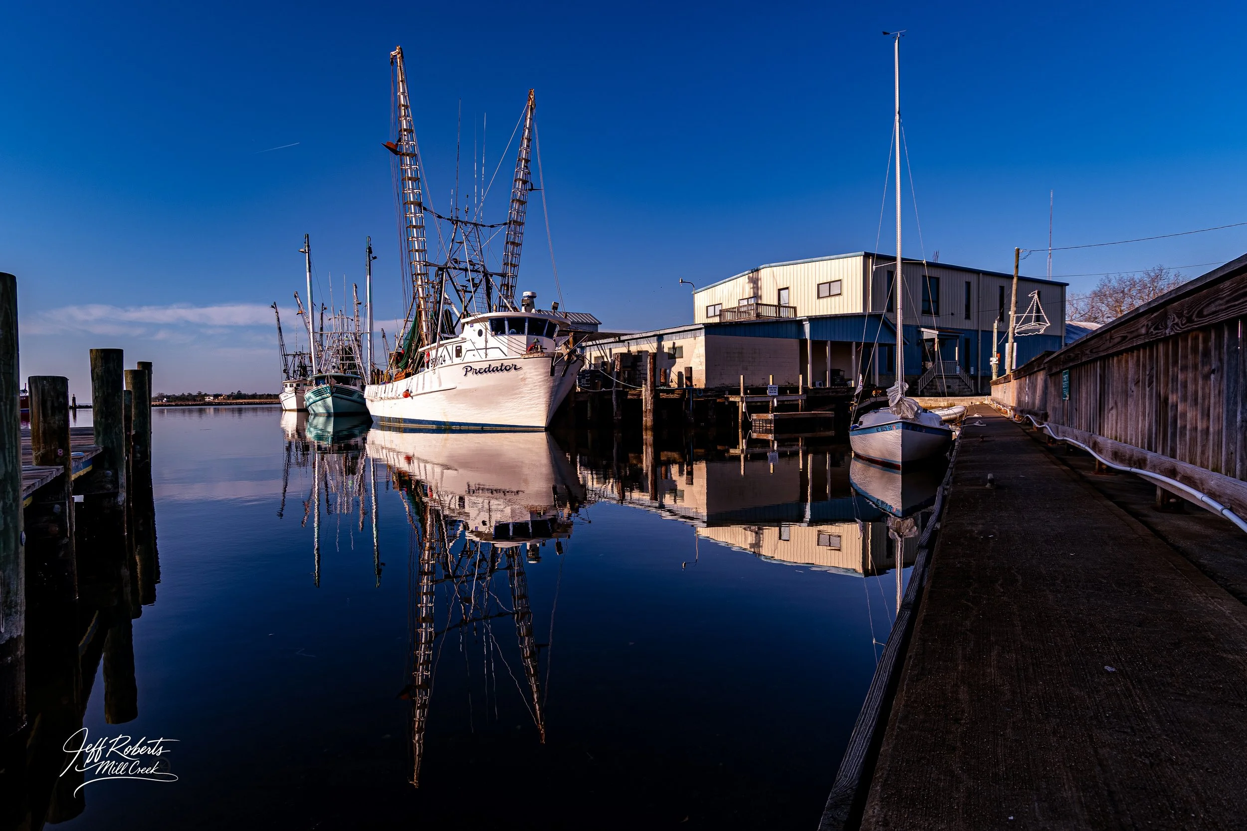 Still water reflection of boats docked at a marina, with buildings and a clear blue sky in the background.