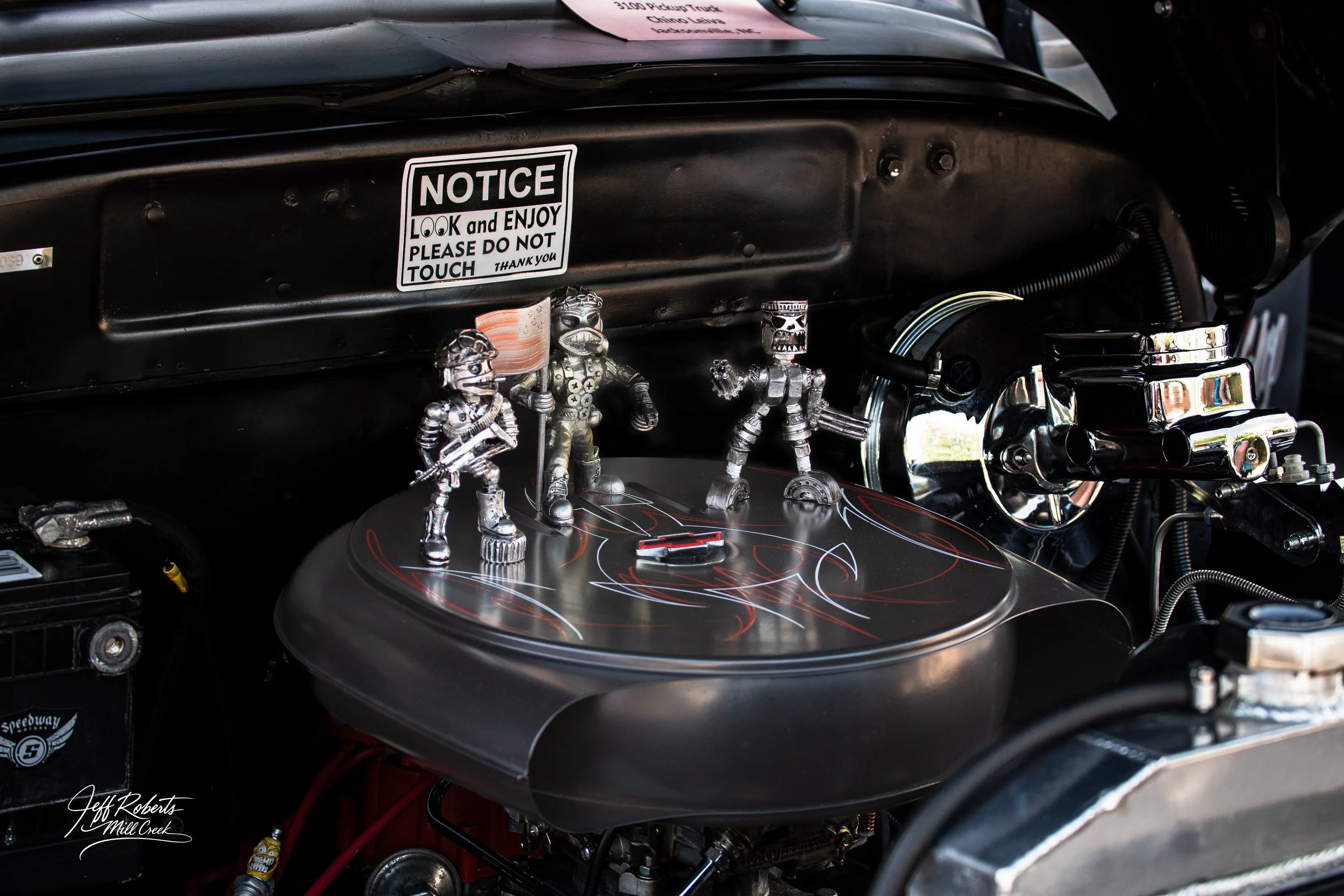 Close-up of a car engine compartment decorated with three small metallic action figures, a sticker that reads 'NOTICE LOOK and ENJOY PLEASE DO NOT TOUCH,' and shiny engine components.