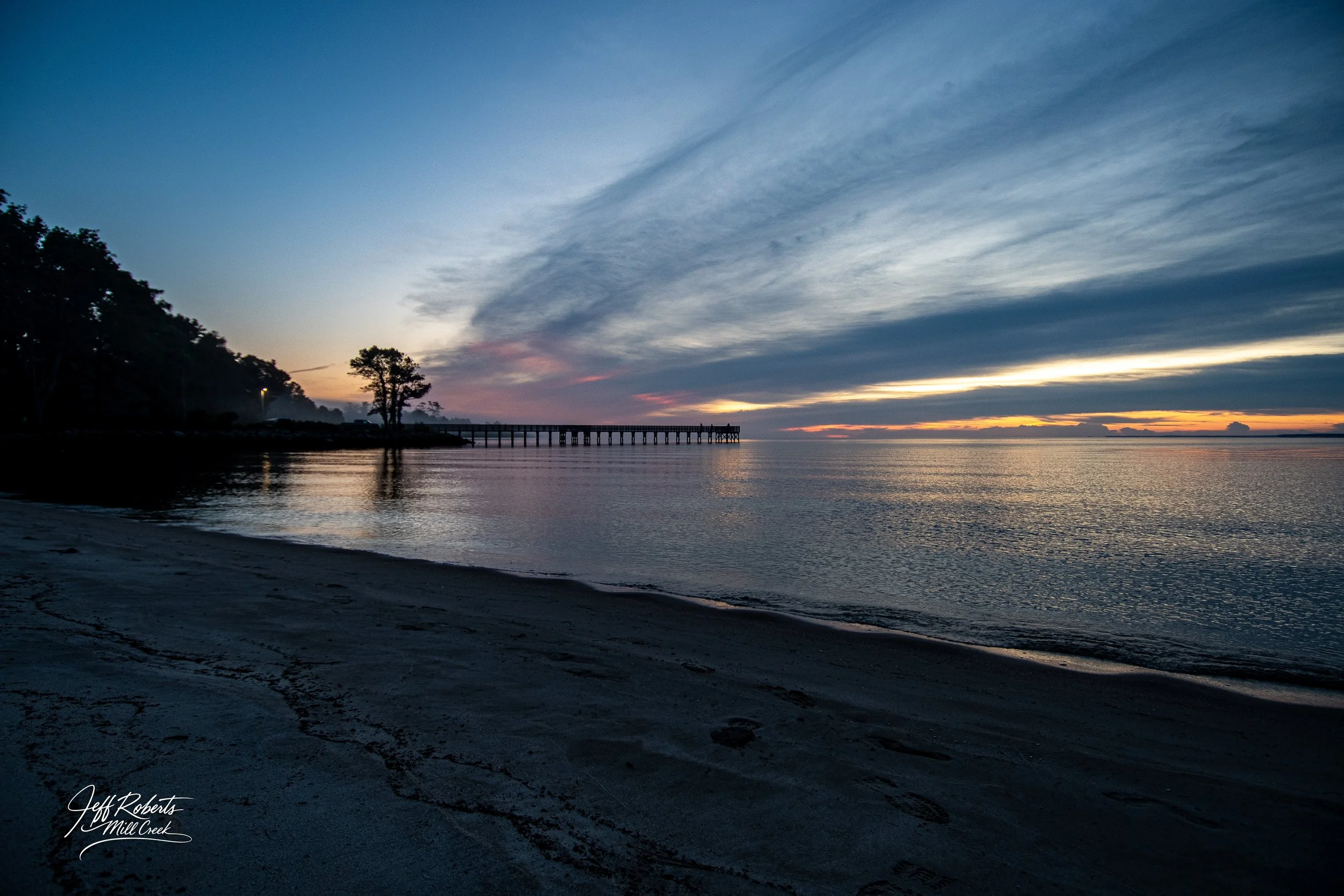 Sunset over a calm beach with a pier extending into the water, silhouette of trees on the left, and a cloudy sky with streaks of orange and pink near the horizon.