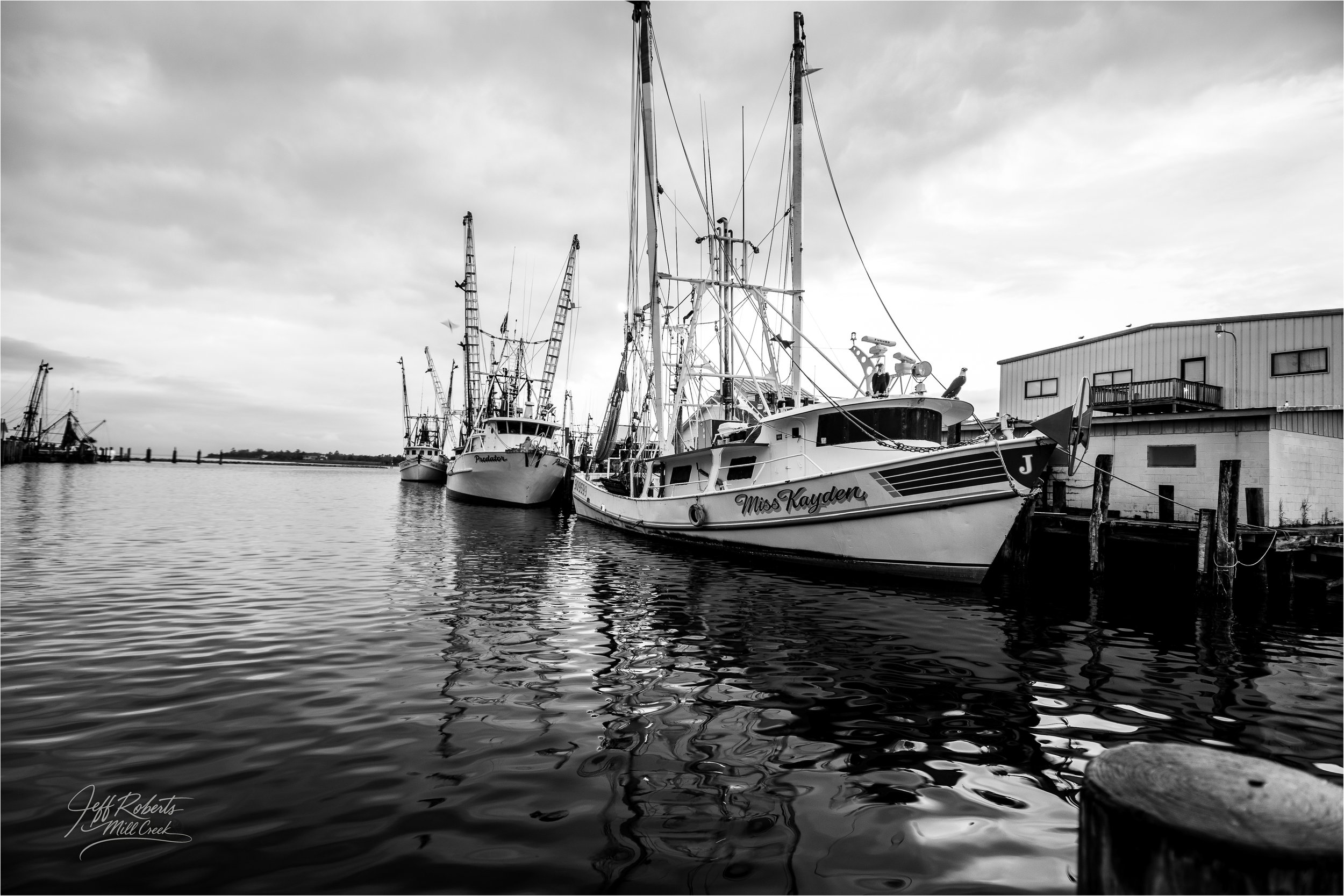 A black and white photo of boats docked at a pier, with a building in the background and the water reflecting the boats and sky.