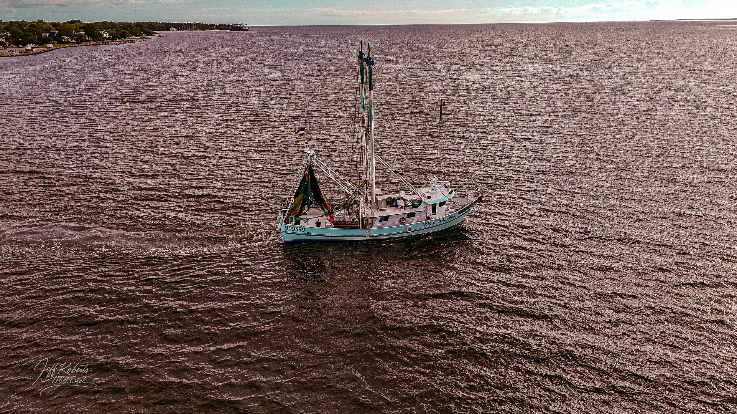 A sailboat on a body of water, with land and trees in the background, seen from above. The sailboat has a white hull and a tall mast.