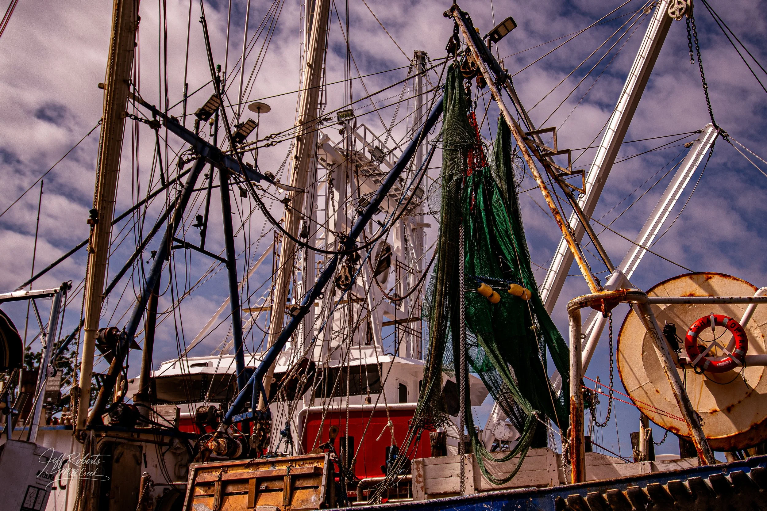 Close-up of a ship's deck with masts, rigging, and nets under a partly cloudy sky.