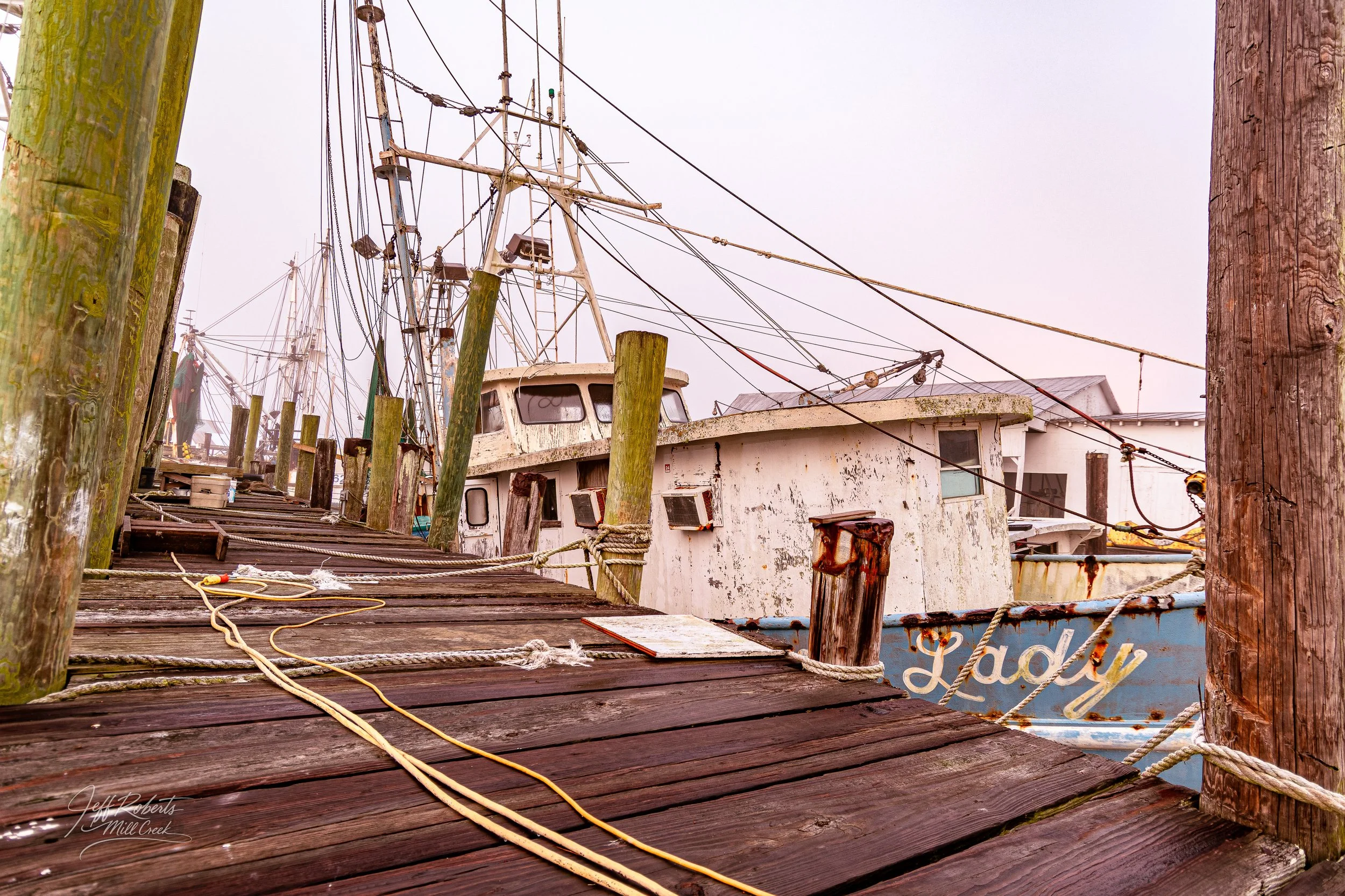 An old, weathered boat docked at a pier with wooden planks, weathered wooden posts, and tangled ropes, surrounded by other boats and a cloudy sky.