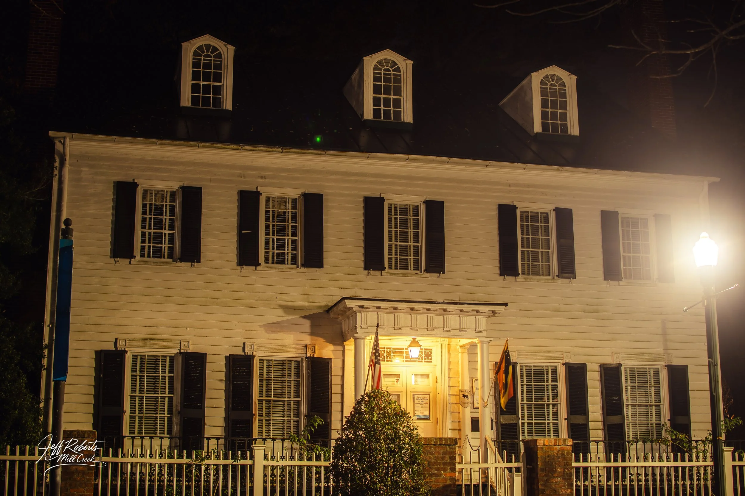 Historic white wooden house with black shutters, illuminated by a streetlamp at night.