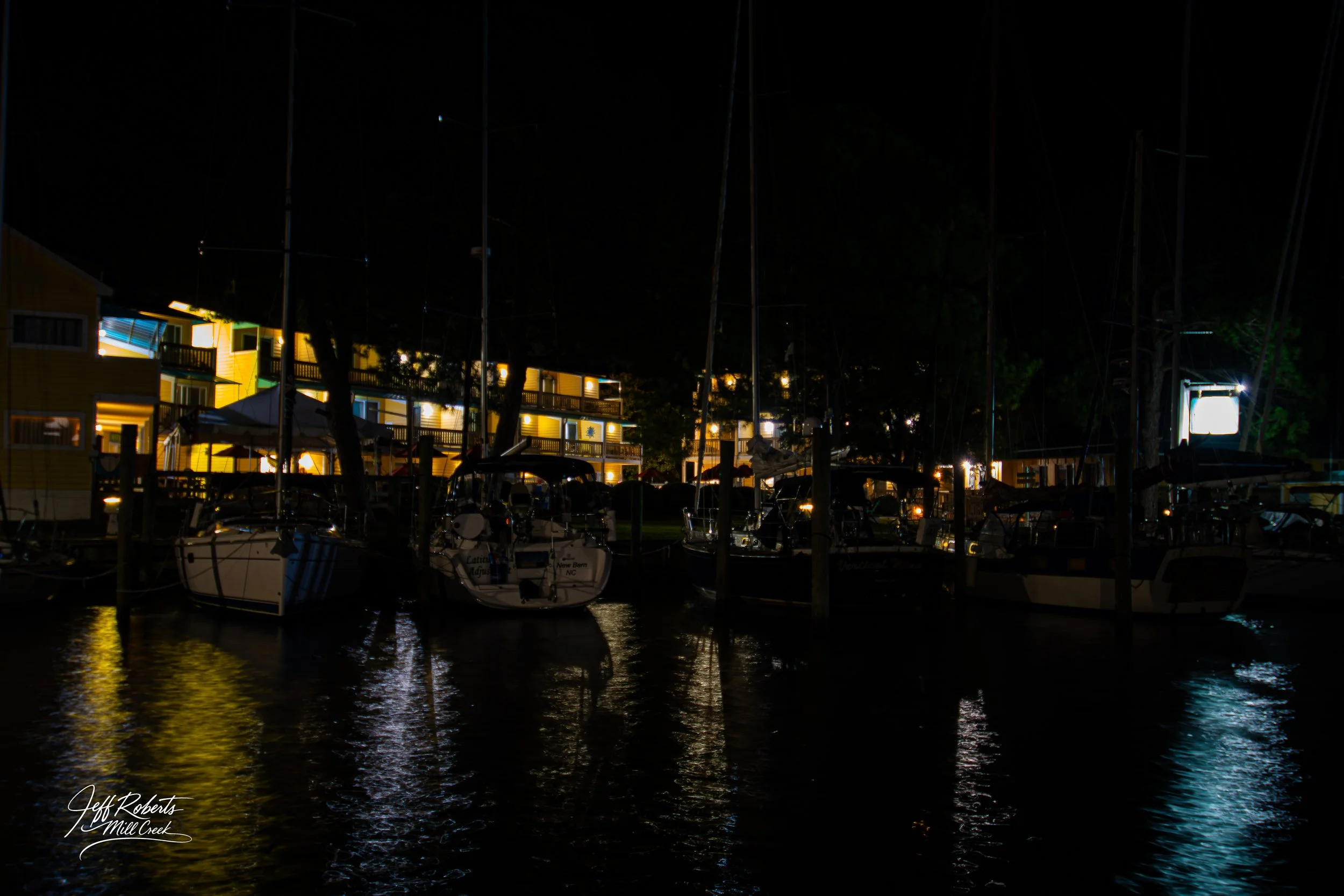 Night view of a marina with boats docked, illuminated buildings in the background, and reflections on the water.