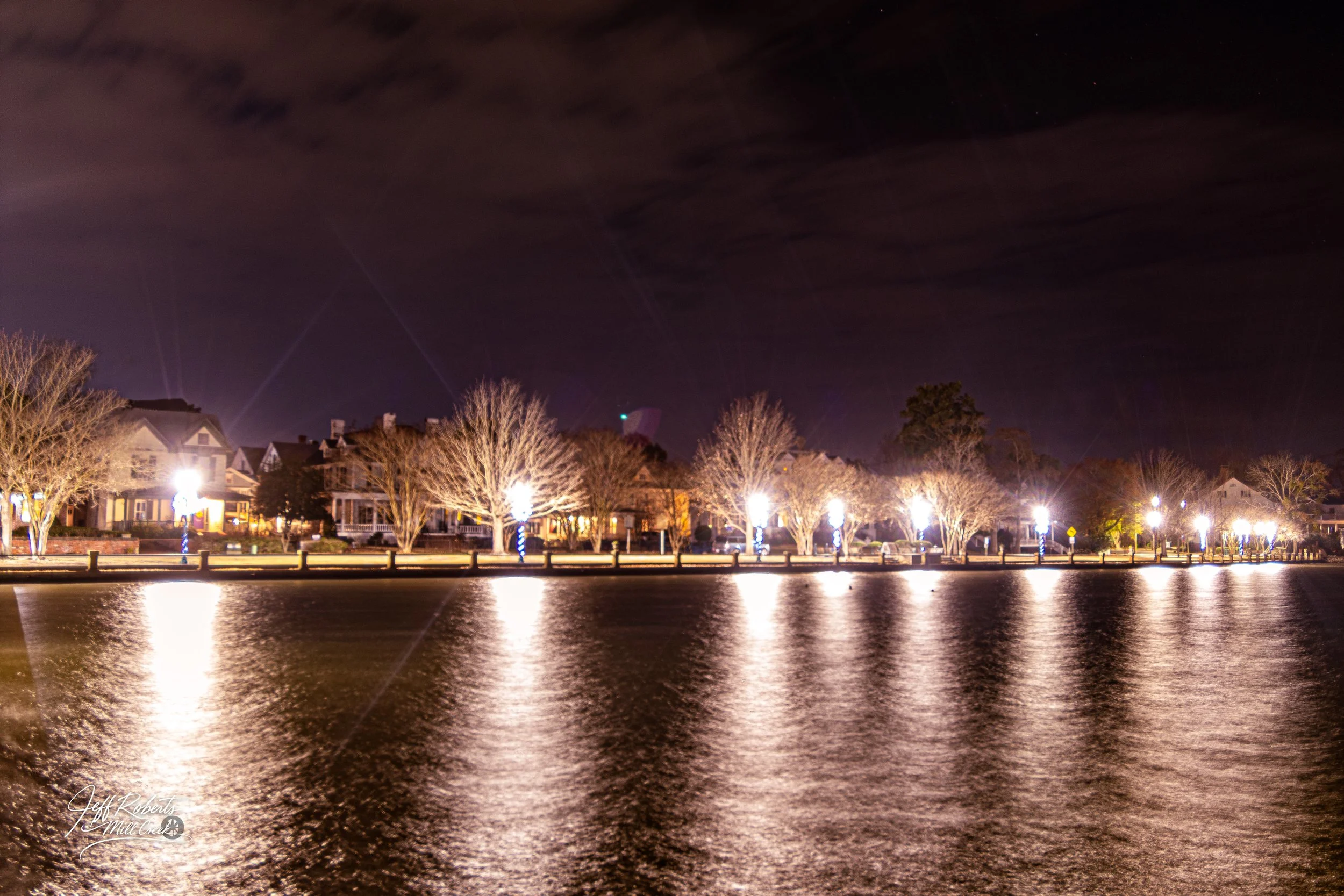 Night view of houses along a waterfront with trees and streetlights, reflected in the water