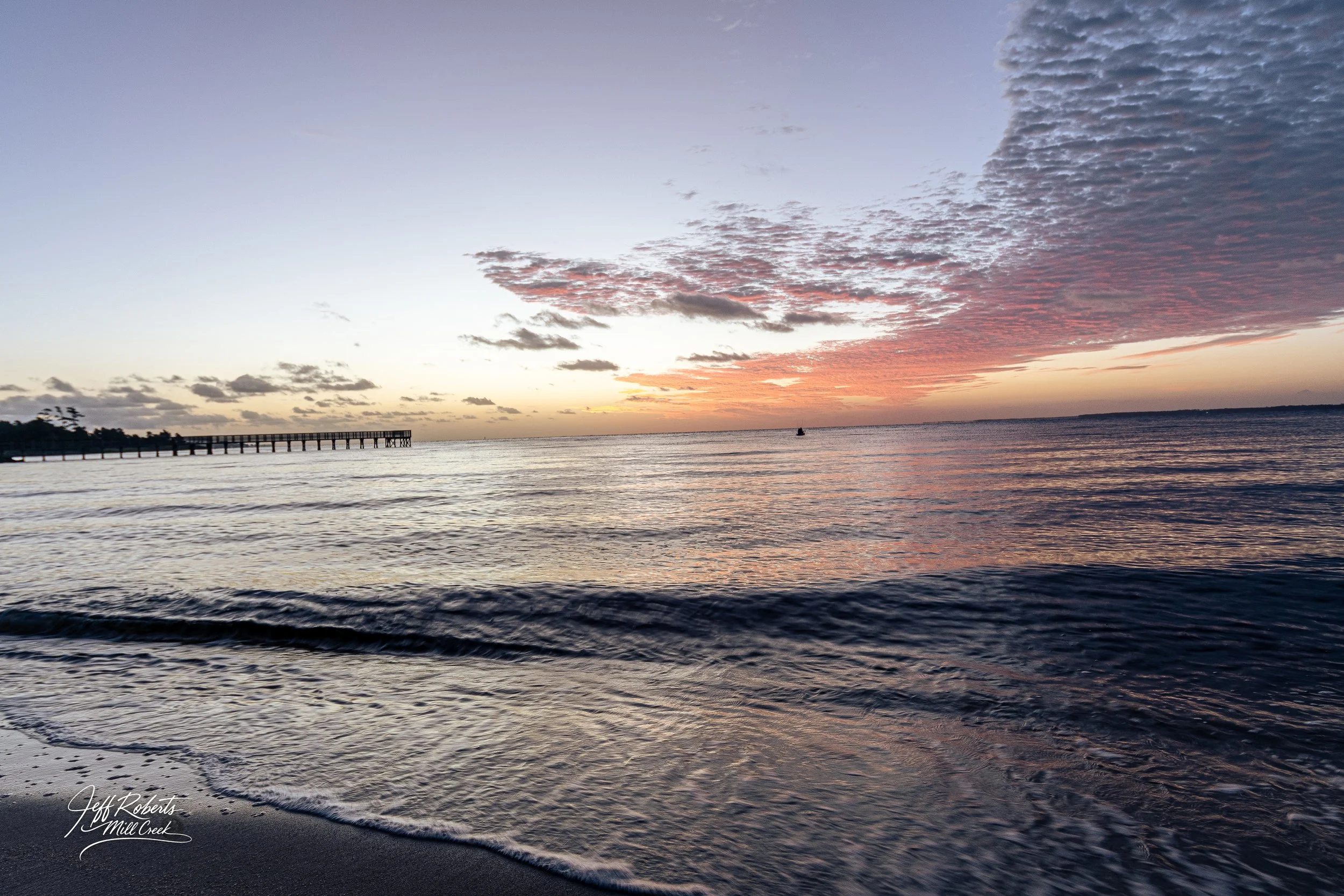 Sunset over a calm ocean with a pier on the left, and a boat near the horizon, sky with clouds illuminated by the setting sun.