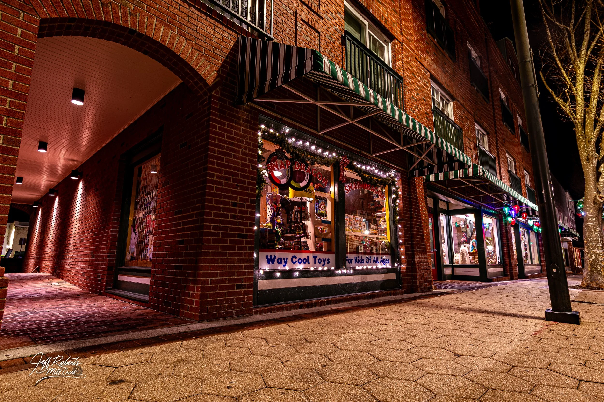 Night view of a toy store called 'Way Cool Toys' with Christmas decorations, lights, and signs, located in a brick building on a city sidewalk.