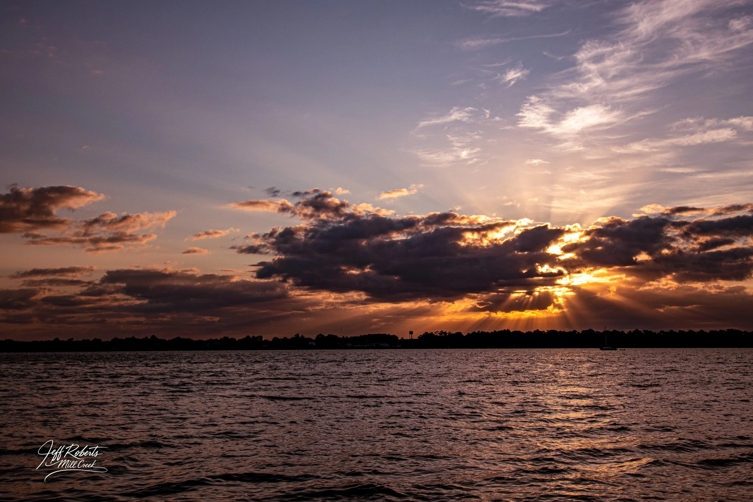 Sunset over a body of water with dark clouds and rays of sunlight breaking through, with a faint sailboat visible on the horizon.