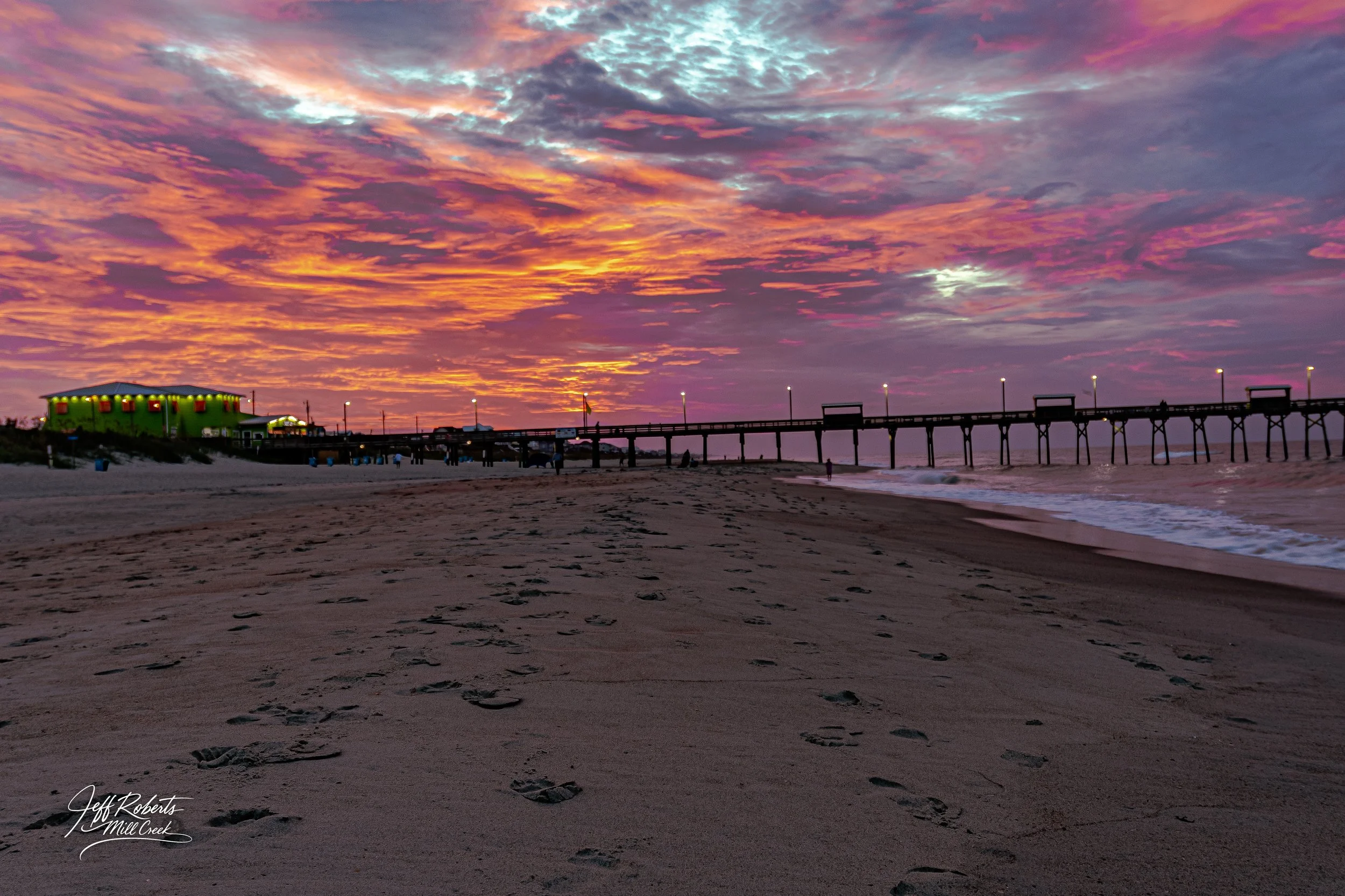Sunset over a sandy beach with footprints, a pier extending into the ocean, and a green building with red lights at the end of the pier.