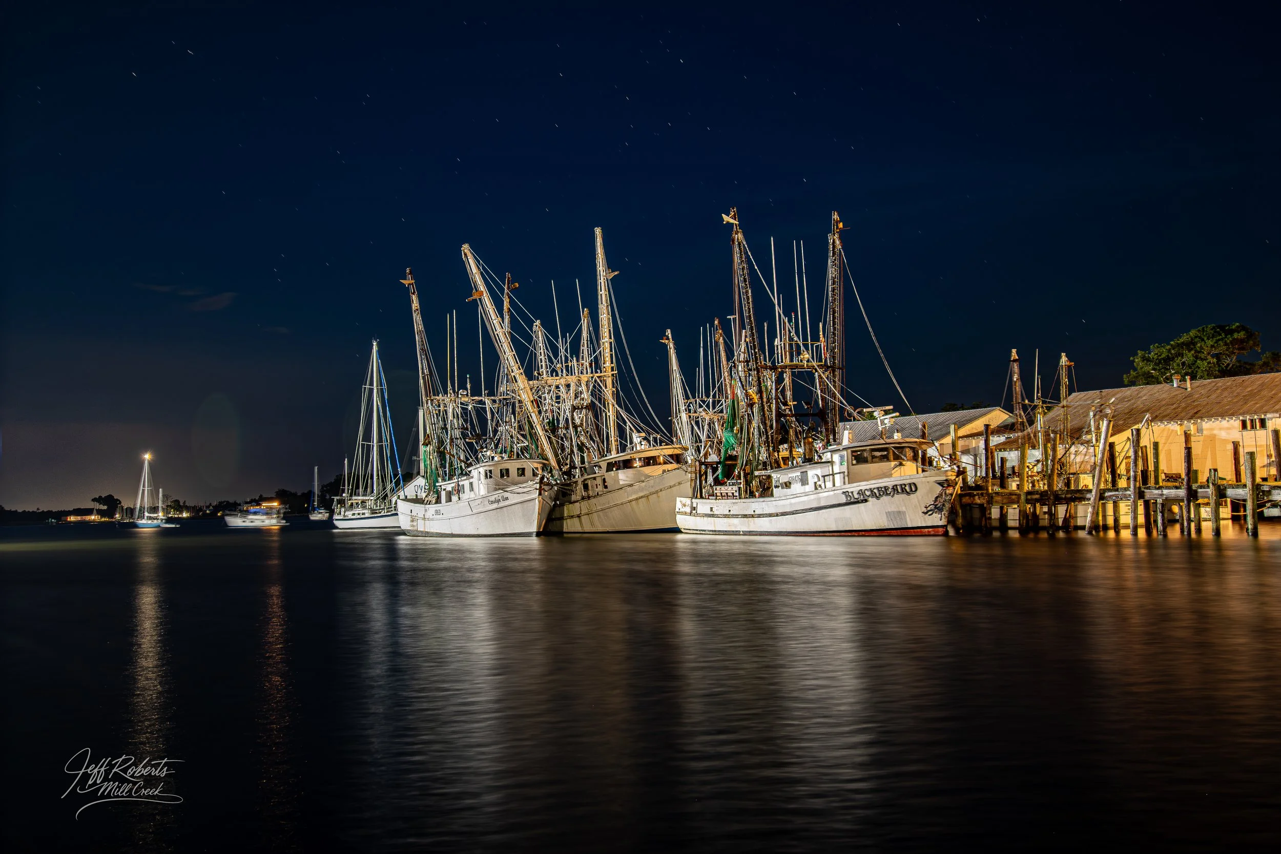 Nighttime view of sailboats and fishing boats docked at a harbor, with reflections on the water and a clear starry sky in the background.