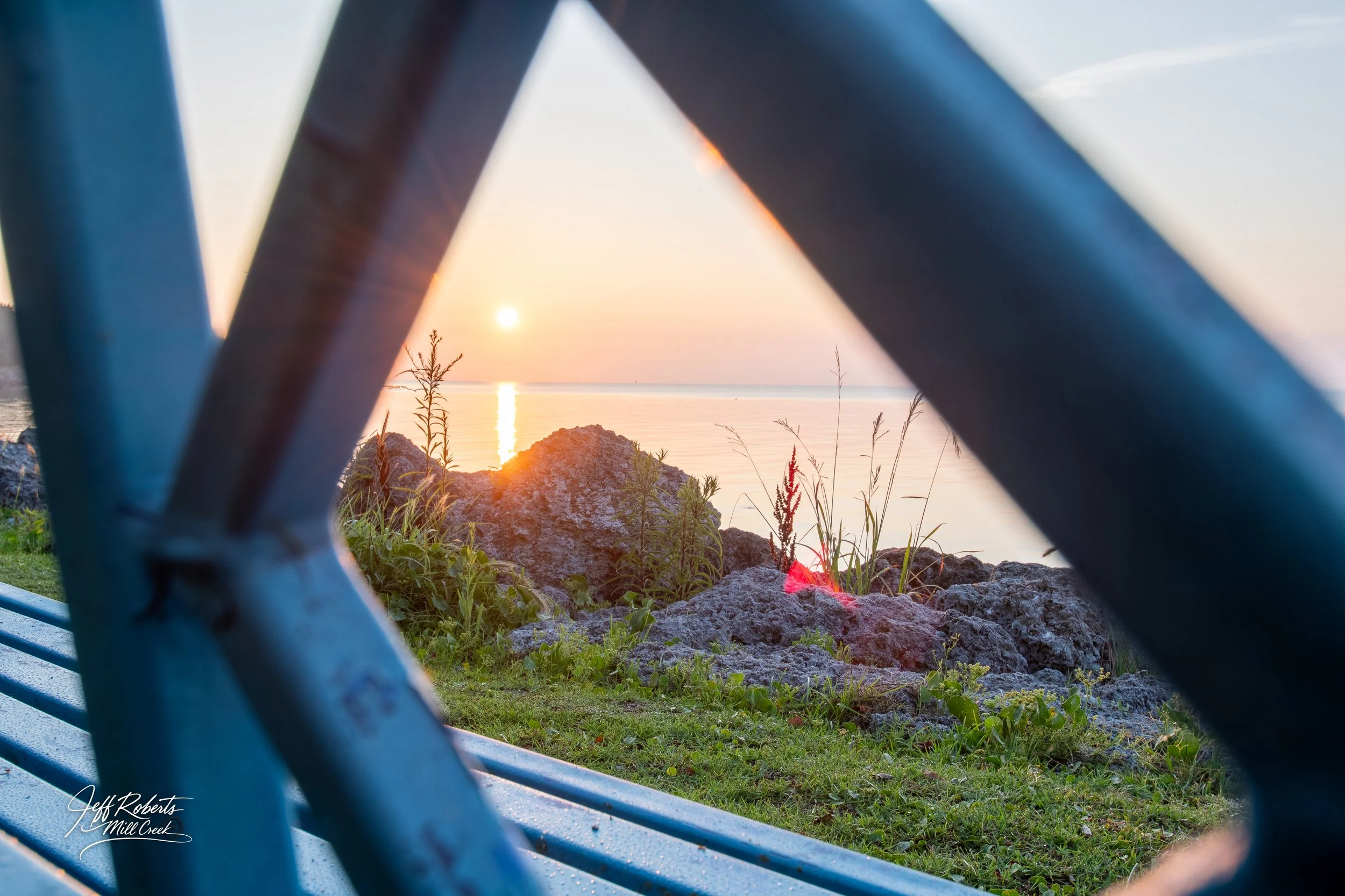 Sunset over a calm body of water, seen through a park bench and metal fence, with rocks and grass in the foreground.