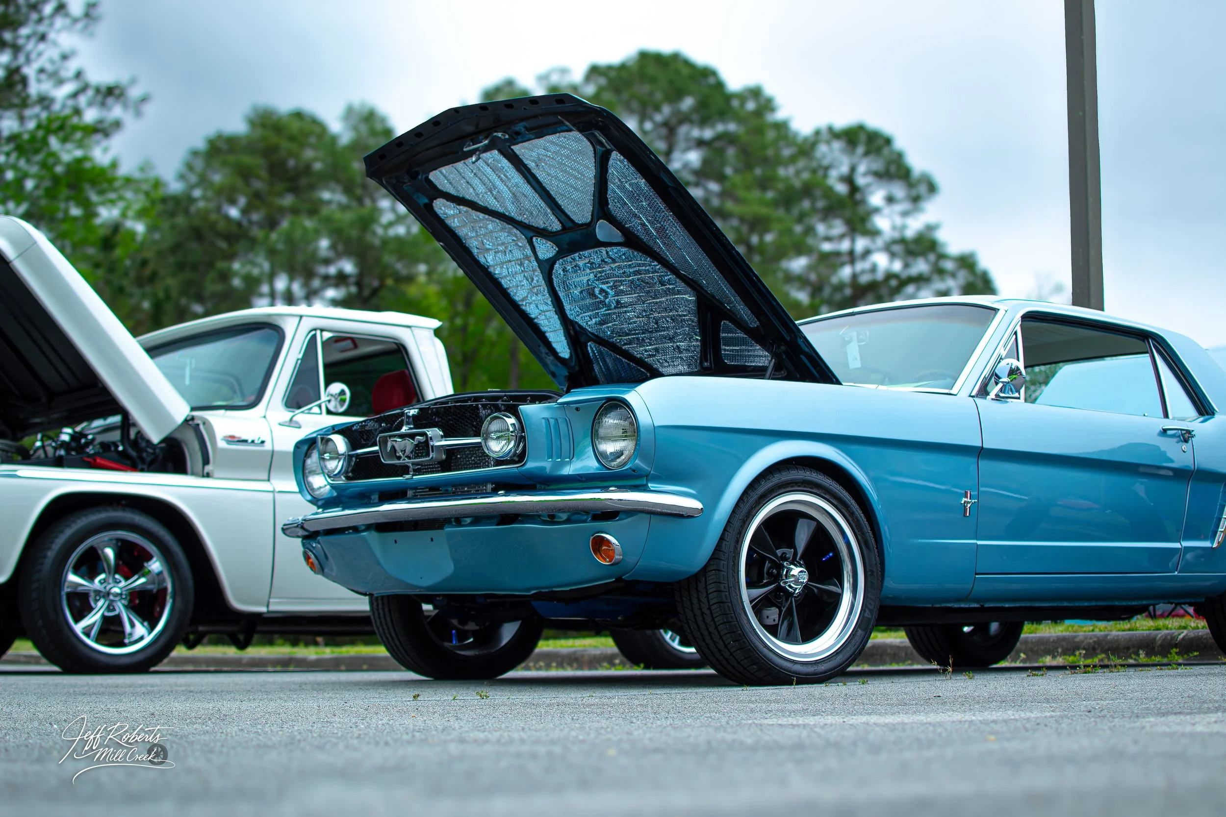 A light blue classic Ford Mustang with its hood open at a car show, parked beside a white vintage truck.