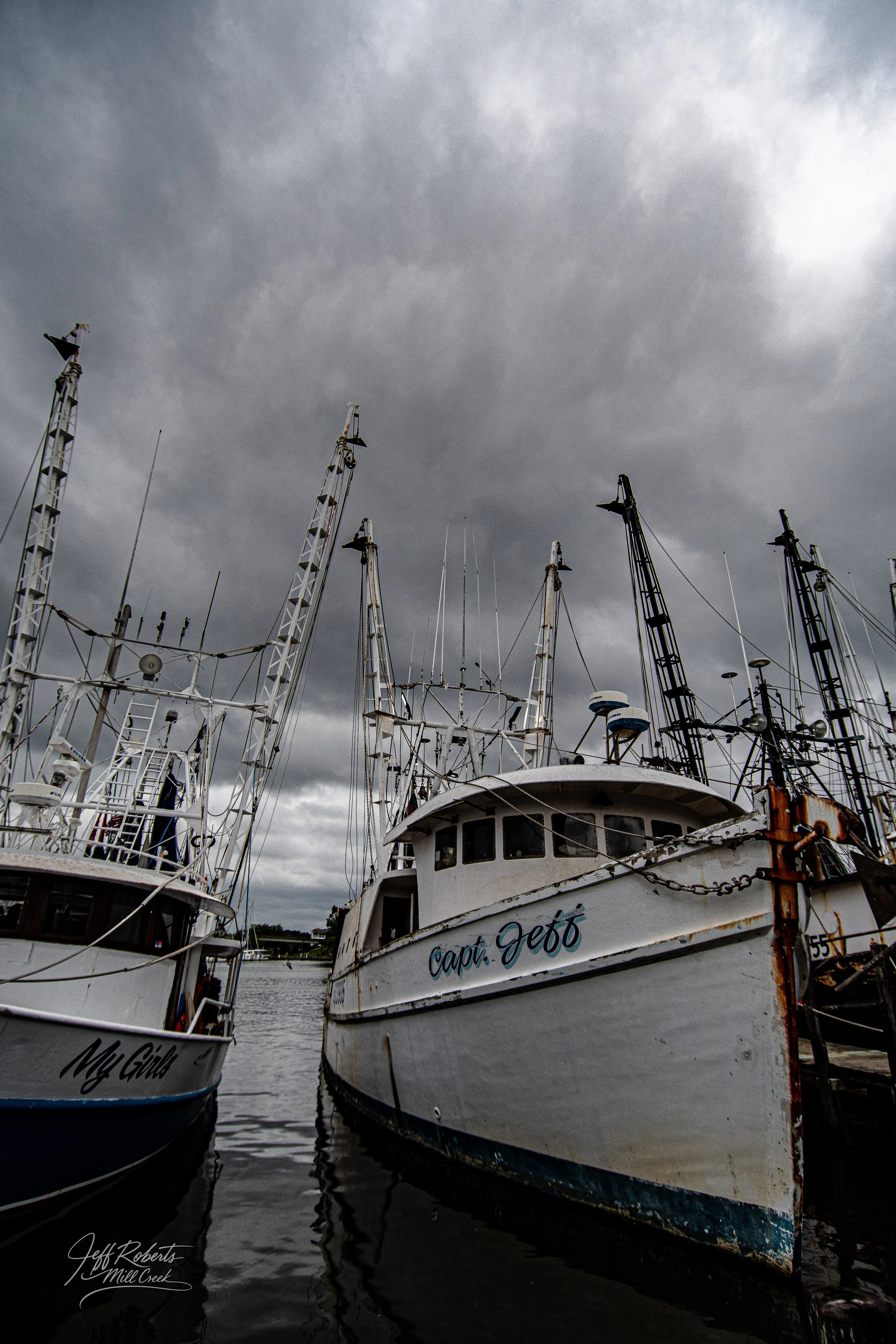 Overcast sky with dark clouds over a dock filled with fishing boats, some showing rust and wear, with their masts rising into the sky.