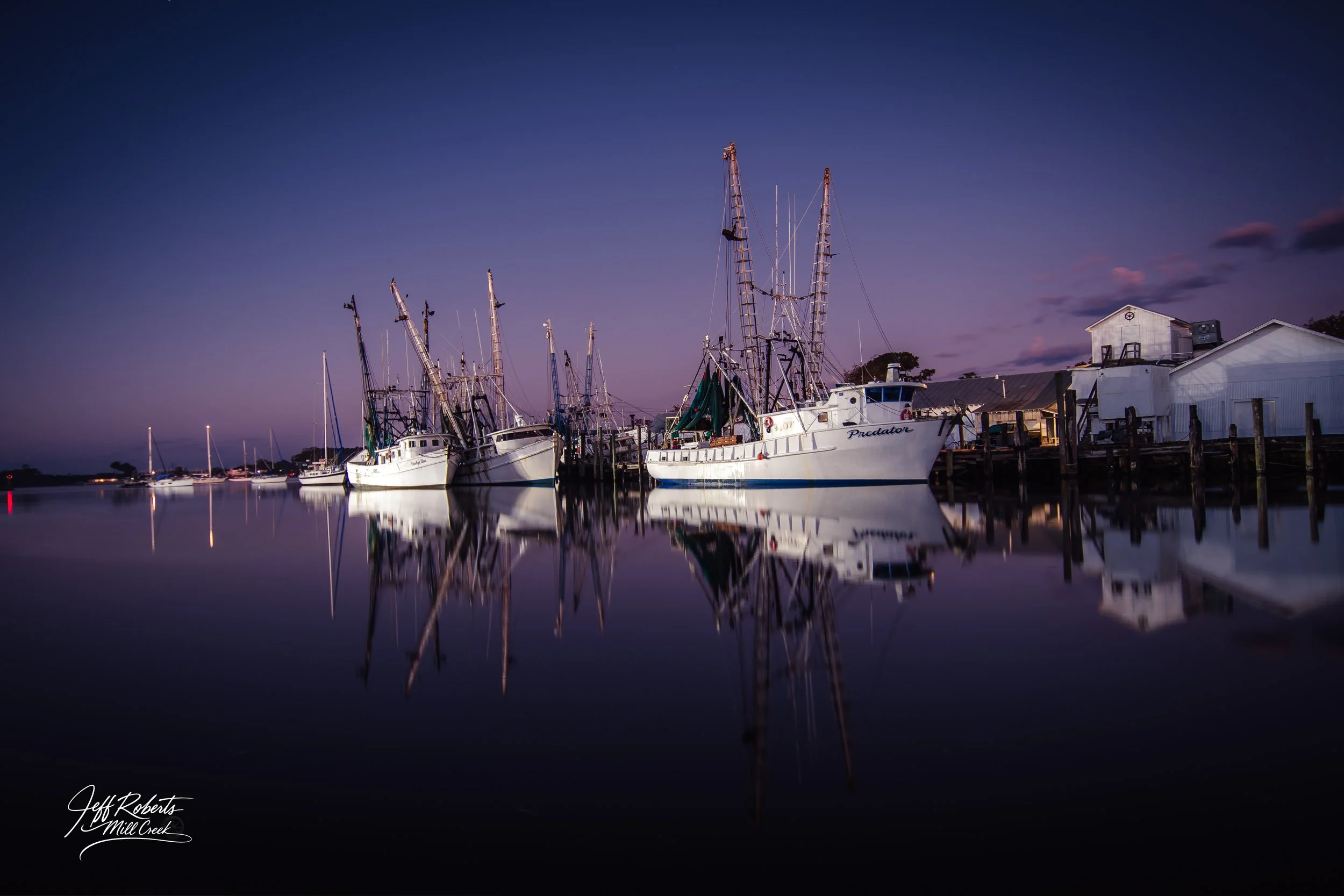 Nighttime view of sailboats docked at a pier with their reflections on calm water, and a few white buildings on the right side.