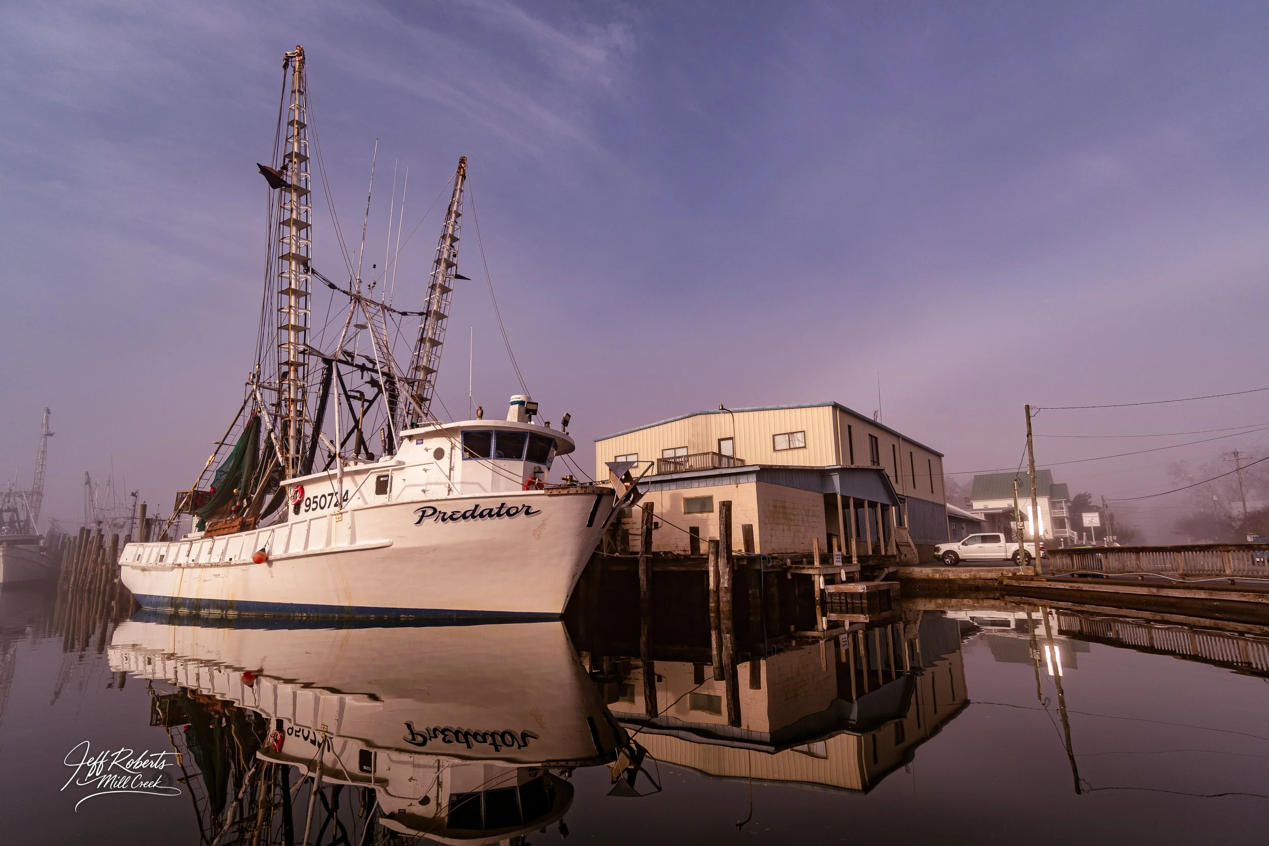 A fishing boat named Predator docked at a marina with its reflection visible in the calm water, with buildings and cars in the background under a foggy sky.