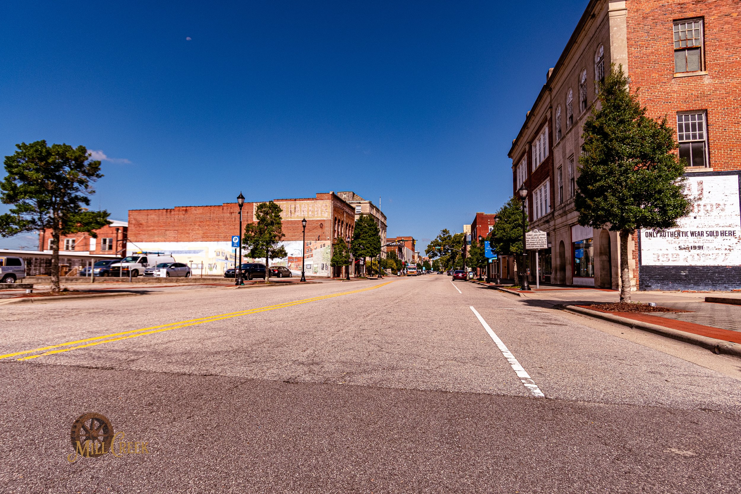 Empty downtown street with parked cars, sidewalks, trees, brick and stone buildings, and a clear blue sky.