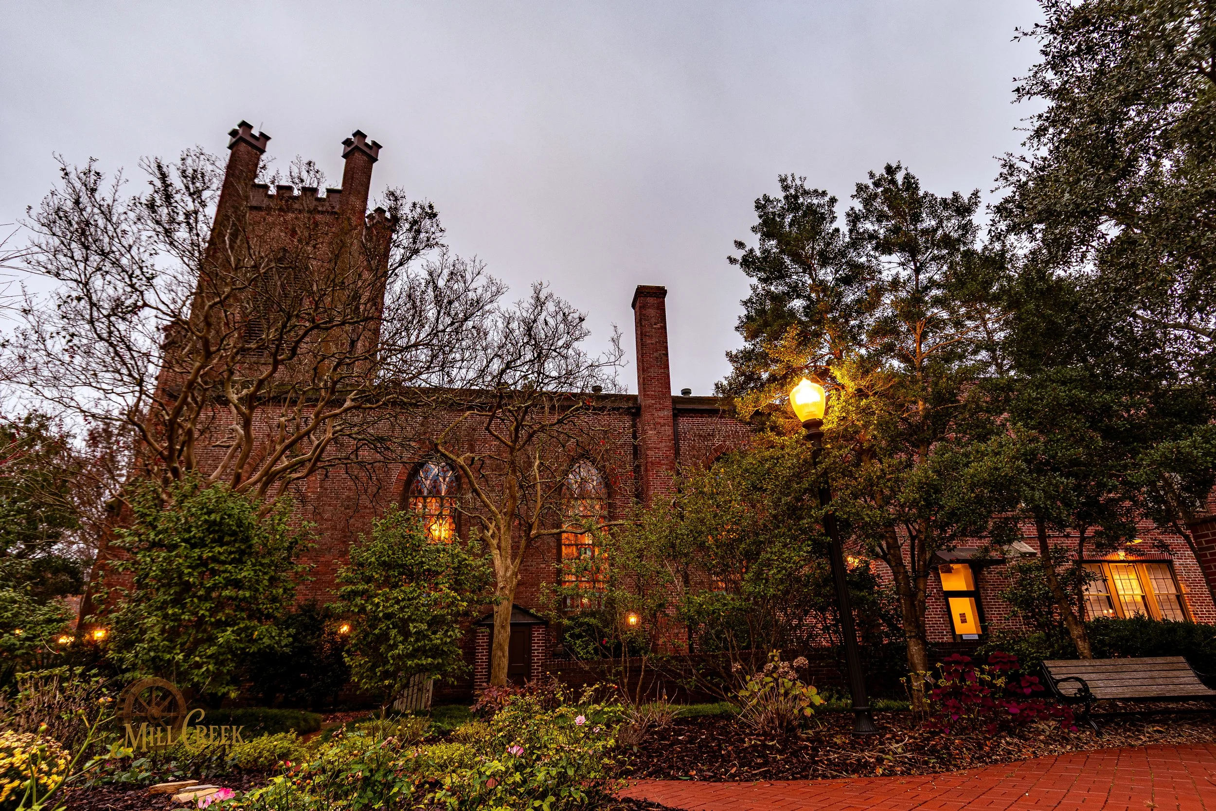 A brick building with a tall tower and arched windows, surrounded by trees and a park with a bench, illuminated by a streetlamp at dusk.