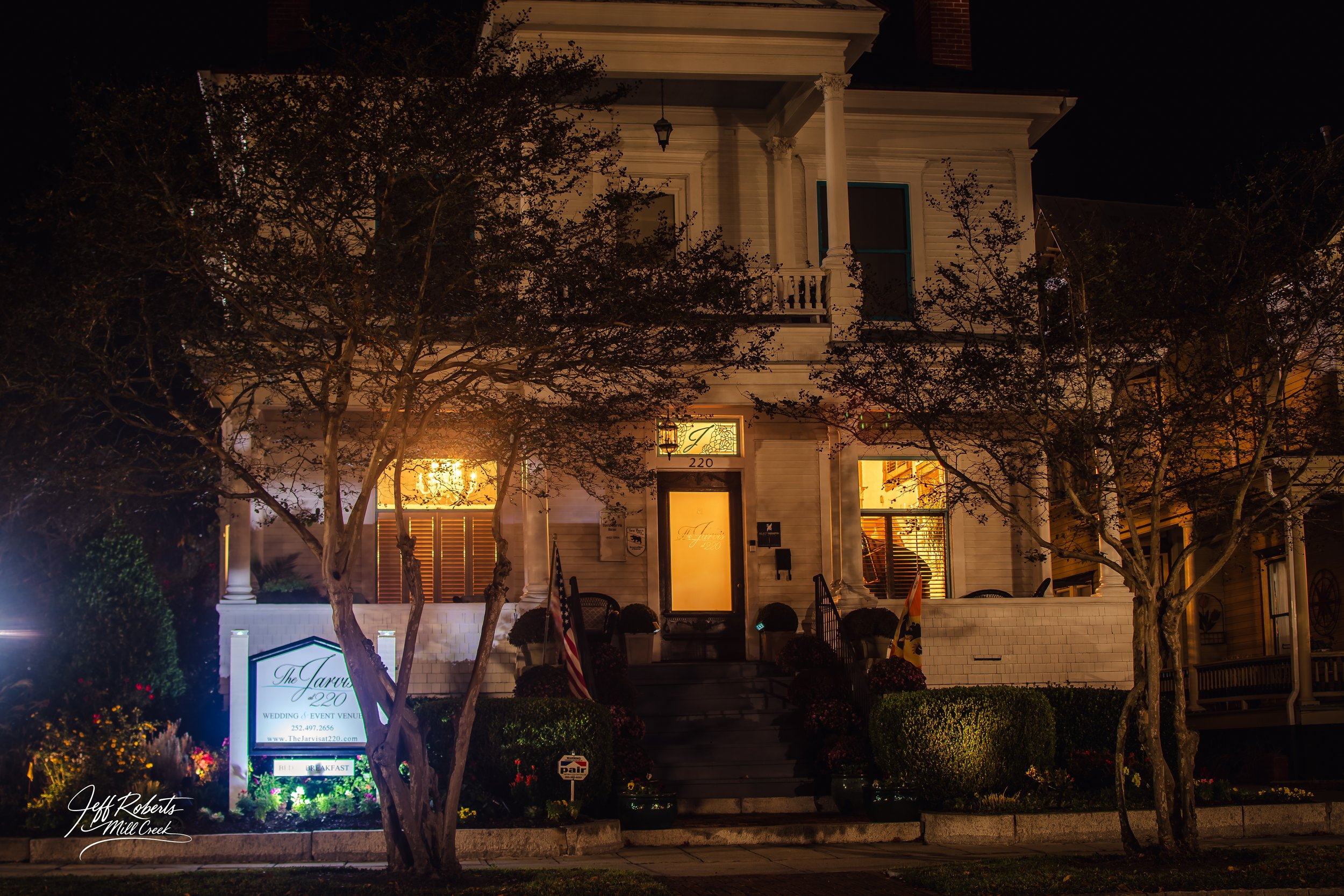 A two-story white Victorian-style house at night with lit interior windows, front porch, and stairs. The house has columns, ornate trim, and a sign in front indicating it is a wedding and event venue, named The Jarvis 220. There are trees, shrubs, an