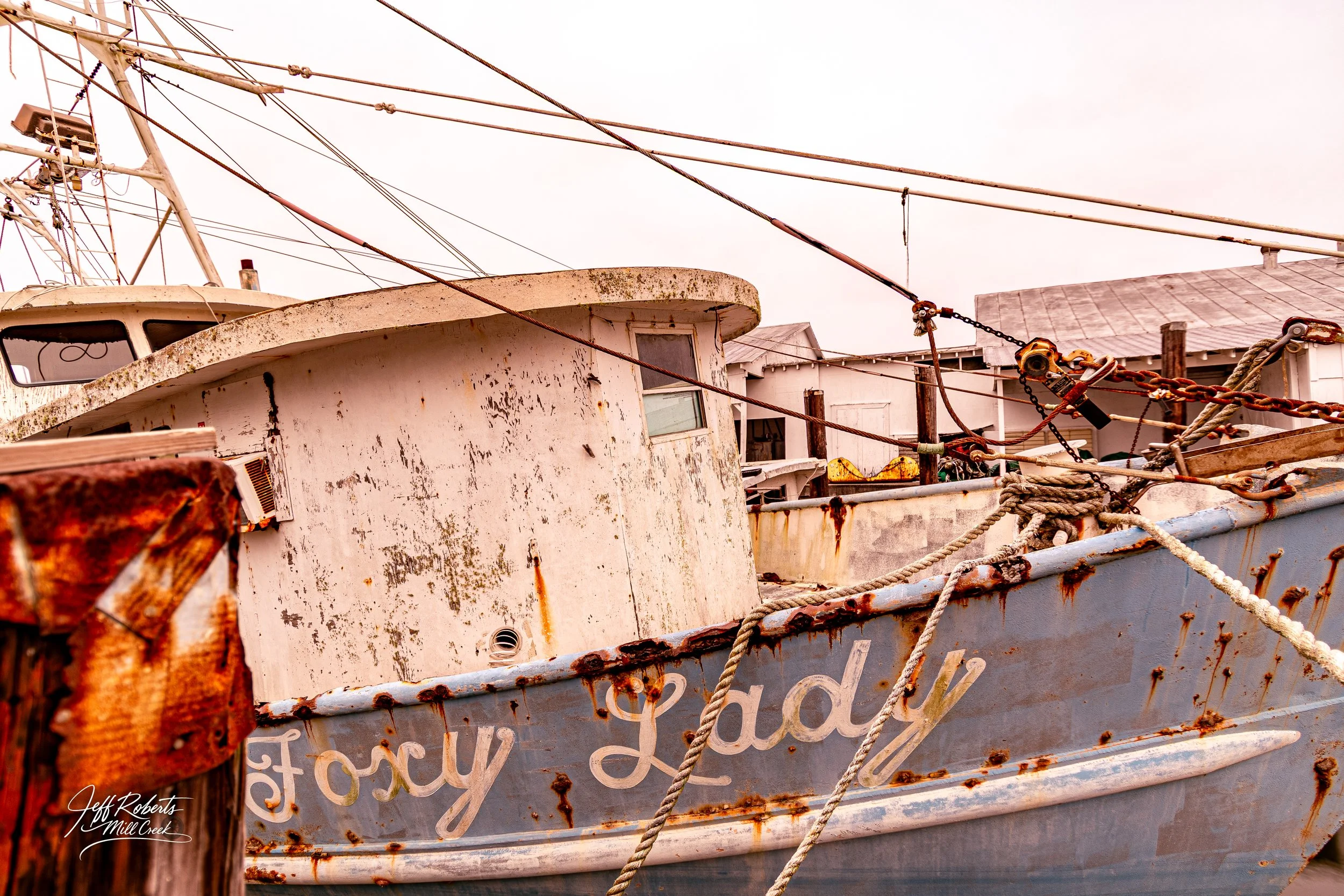 An old, rusty boat named 'Foxy Lady' moored at a dock with weathered ropes tied to wooden posts. The boat's surface is covered with peeling paint and rust stains. In the background, there are small white buildings and a cloudy sky.