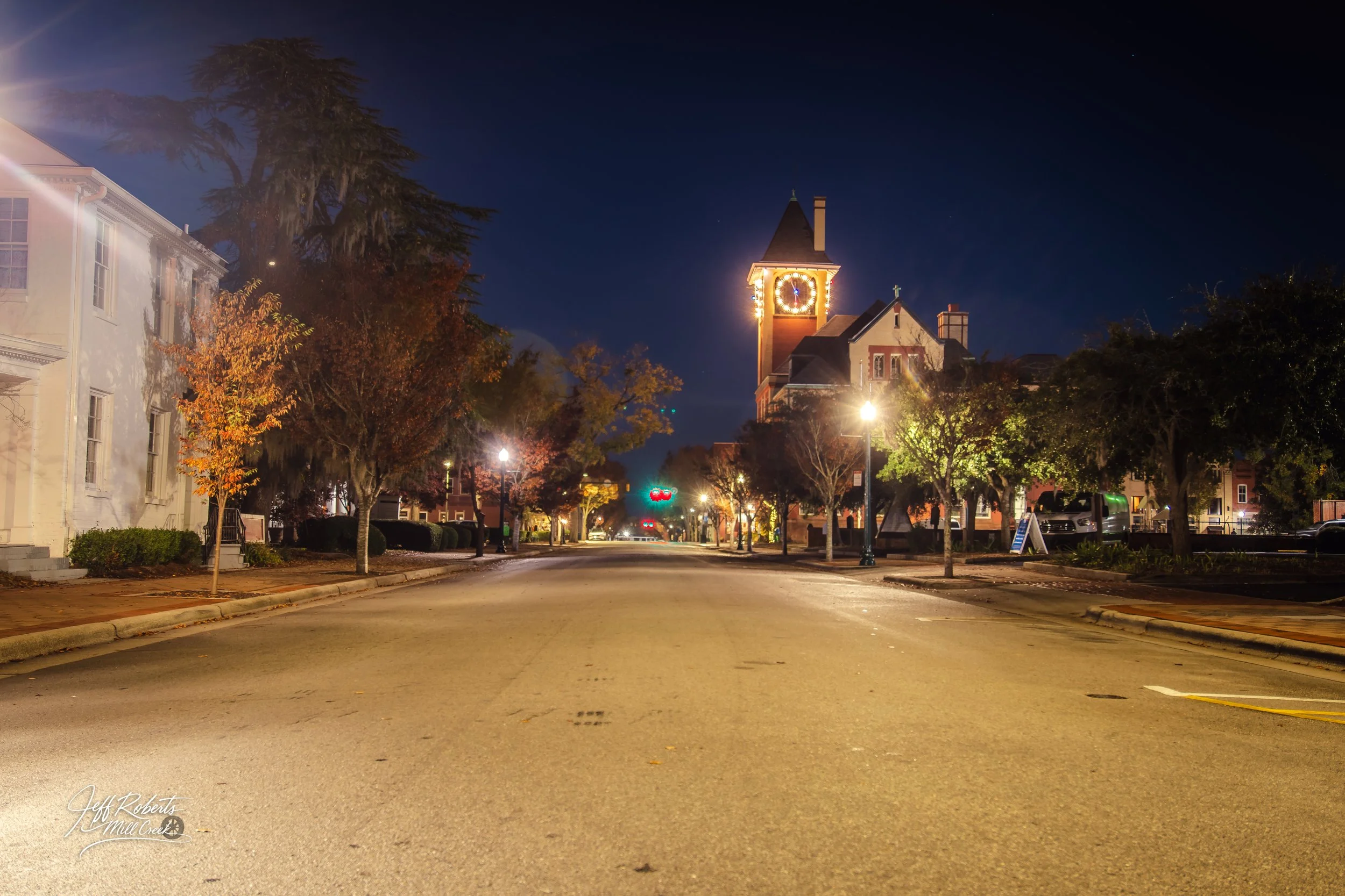 Empty street in a small town at night with illuminated buildings and streetlights, including a clock tower in the background.
