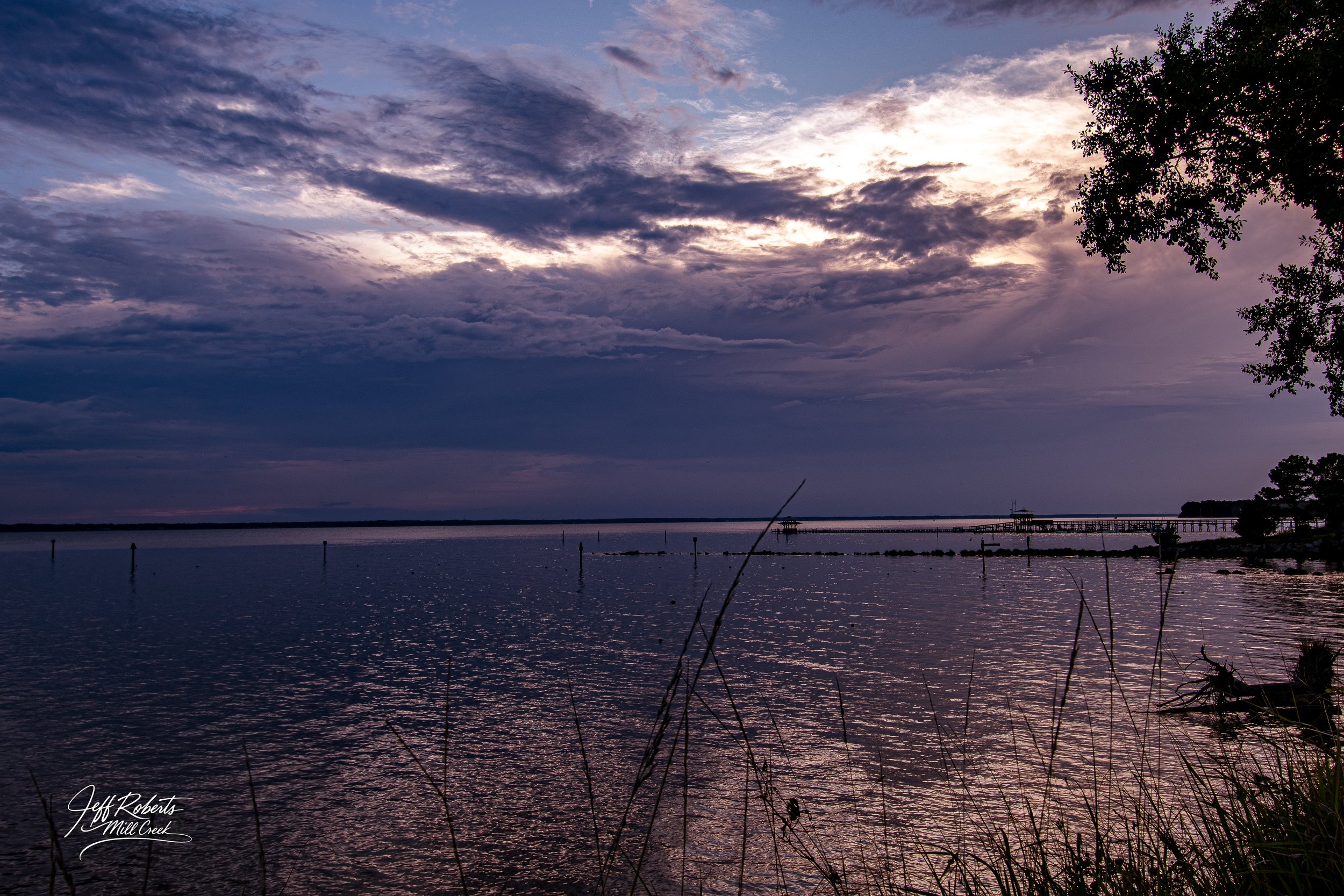 Sunset over a calm body of water with clouds in the sky, trees on the right, and a pier extending into the water.