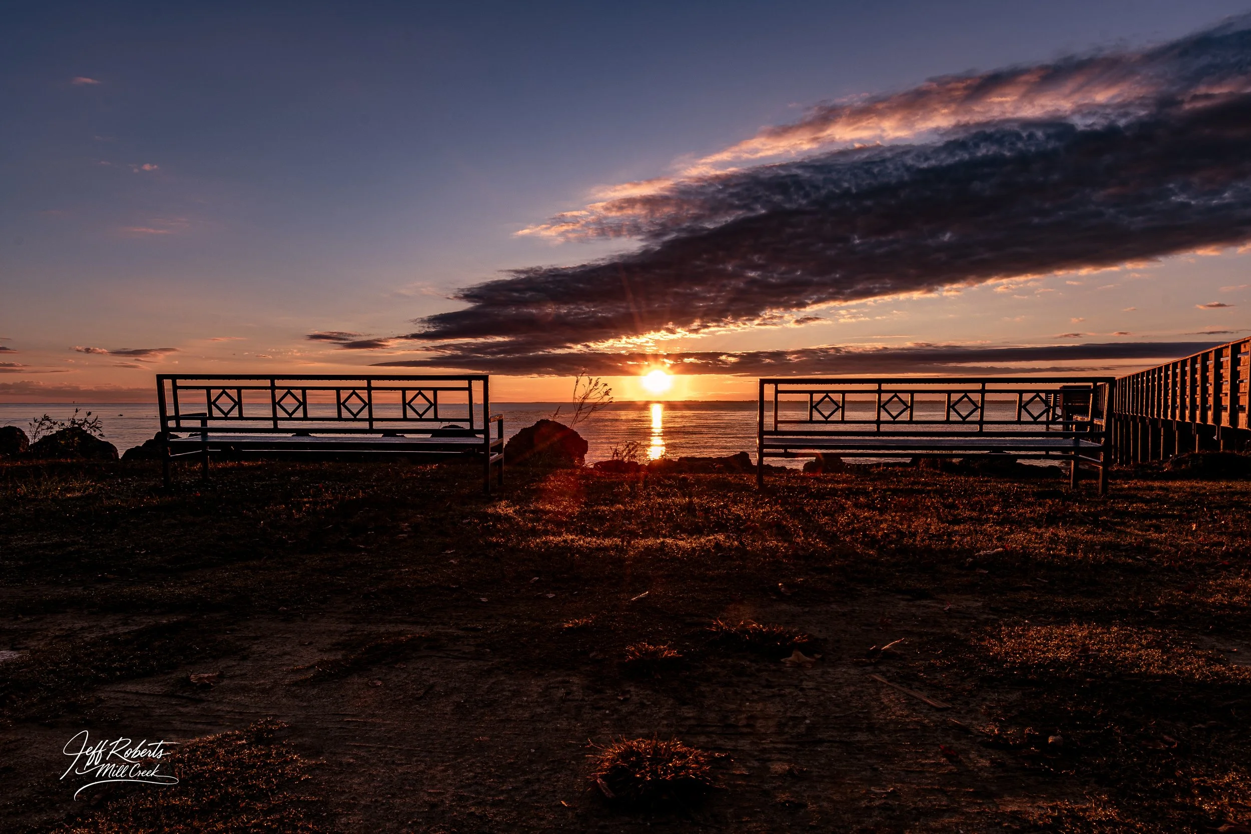 Sunset over the ocean with clouds, two empty benches facing the water, and a wooden walkway on the right side.