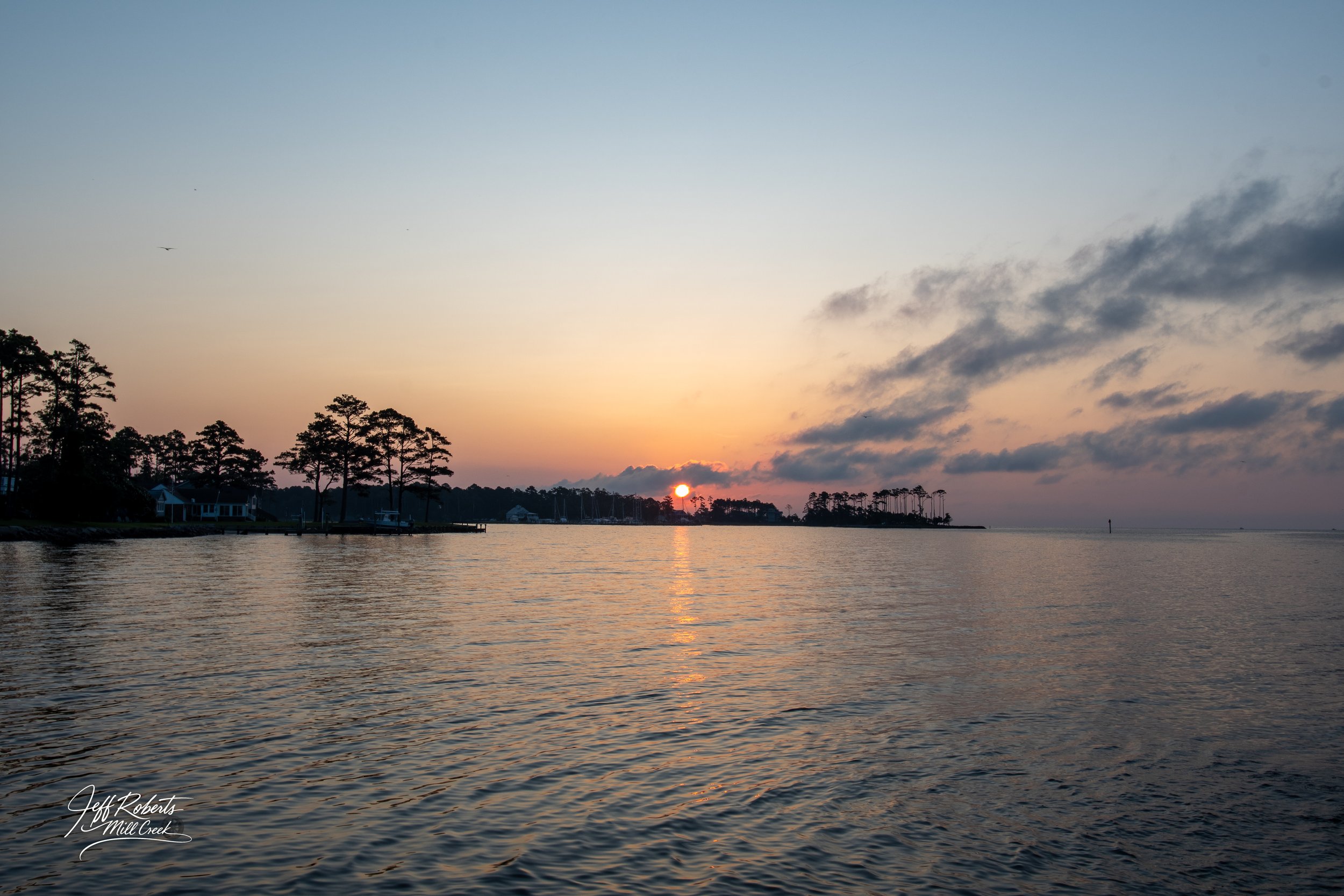 Sunset over a calm body of water with silhouetted trees along the shoreline and a cloudy sky.
