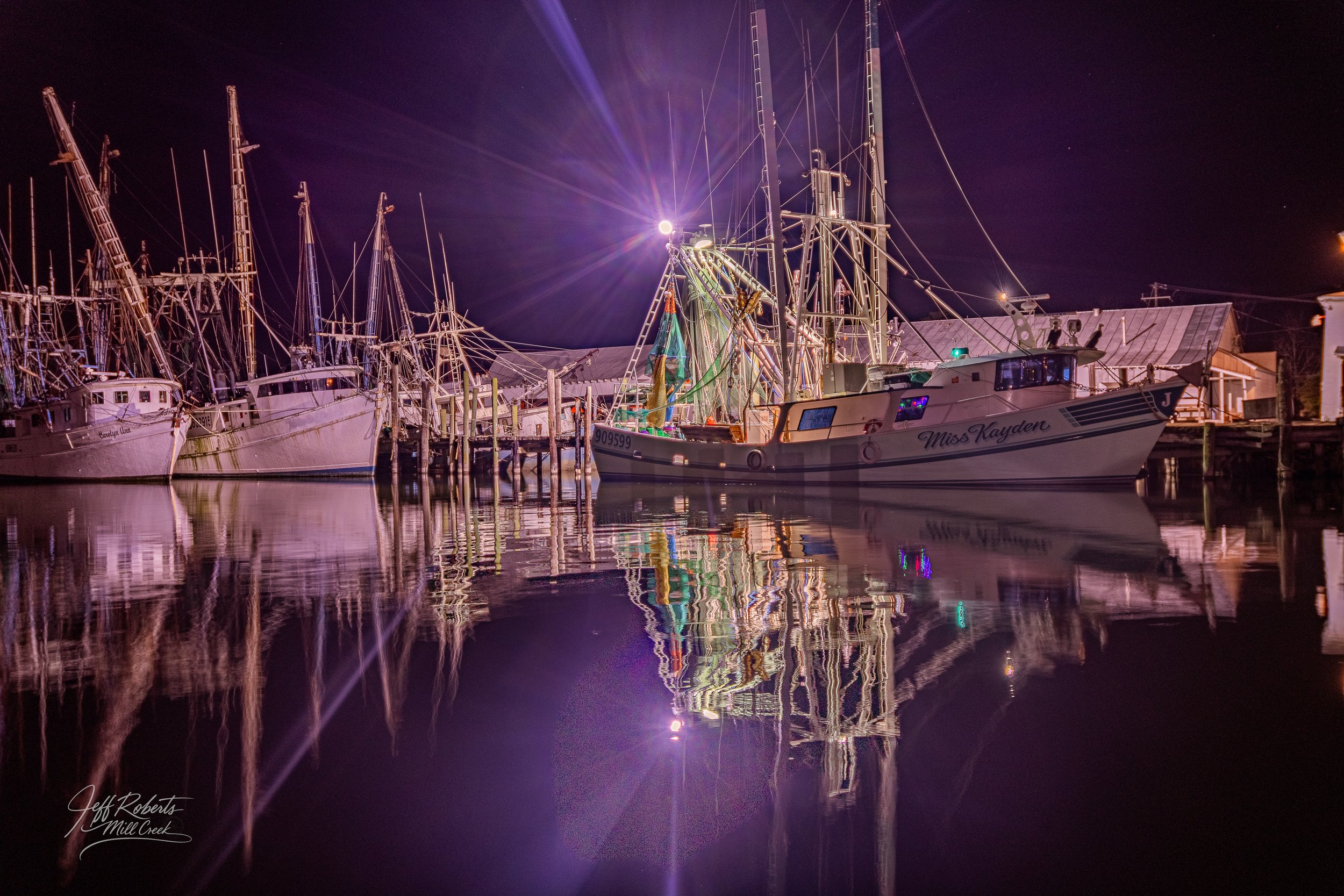 Nighttime scene of a marina with sailboats and a motorboat named Miss Kayden, reflected in calm water, illuminated by a bright light on the boat with purple and pink hues in the sky.