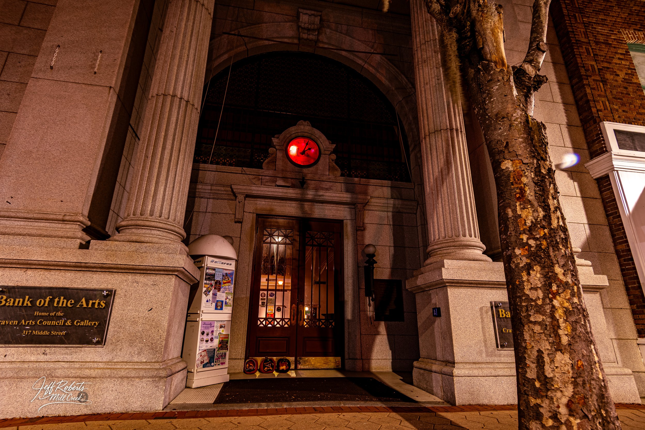 Night view of the entrance to the Bank of the Arts building with stone pillars, a red clock, a tree in the foreground, and Halloween decorations at the door.